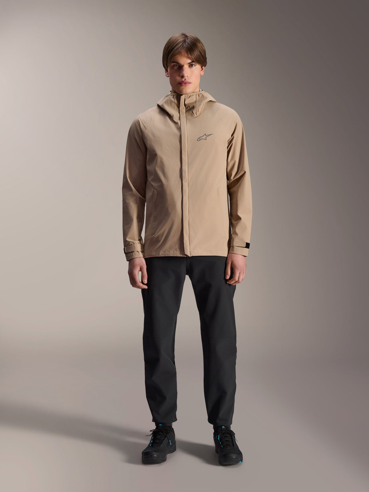 Alpinestars A-Dura Rain Jacket, Light Brown cycling jacket, worn with black pants and black shoes, featuring a small chest logo and hood, professional mountain bike apparel for all-weather riding.