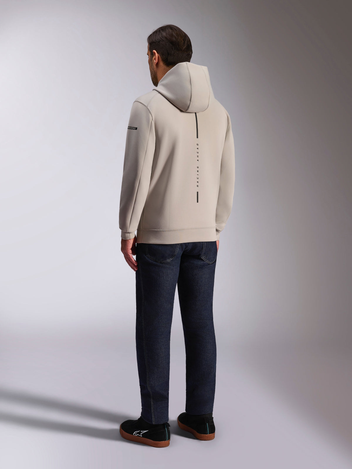 Alpinestars Insight Hoodie, back view in cement gray, features a vertical black stripe with 'RACING AHEAD' text, casual streetwear design, worn with dark jeans and black shoes.