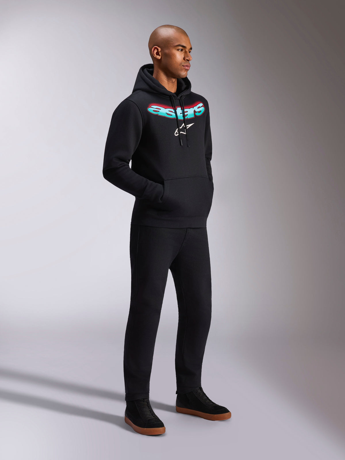 Alpinestars Ellipsoid Hoodie, Black Hooded Pullover Fleece, featuring a curved teal and red gradient Astars wordmark and white star logo, worn by a model with black pants, casual motorsport-inspired streetwear