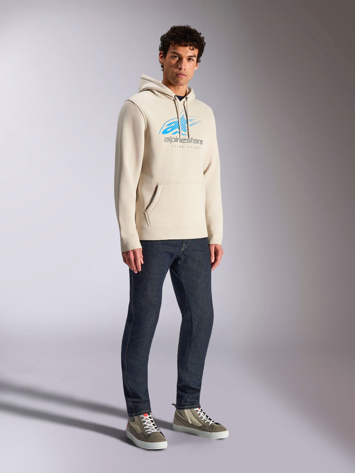 Alpinestars Pulse Hoodie, Off White casual streetwear, hooded pullover fleece featuring a blue and grey Astars logo with speed line fade, kangaroo pocket, worn by model with dark denim jeans and high-top sneakers.