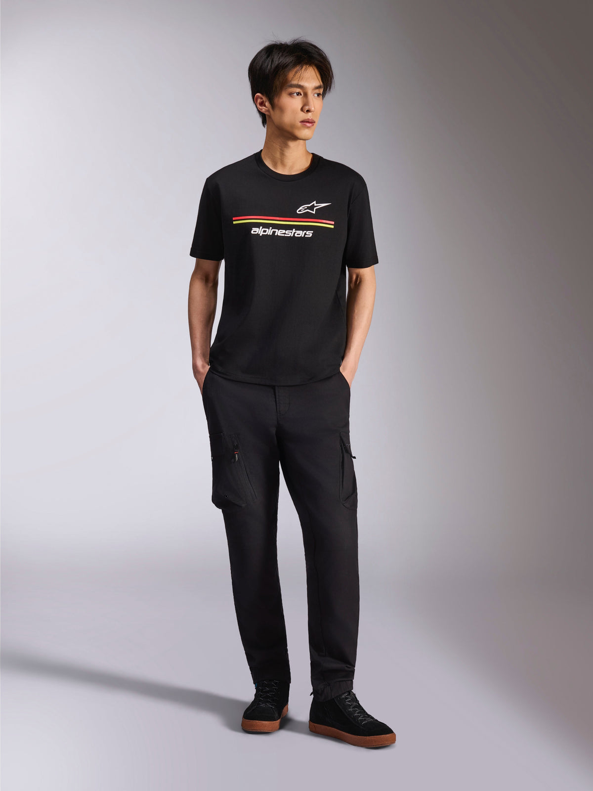 Model wearing Alpinestars Lanes CSF Tee, Short Sleeve T-Shirt, Black, featuring red and yellow stripes with Astars and Alpinestars logos, paired with black cargo pants and sneakers, casual motorsport streetwear