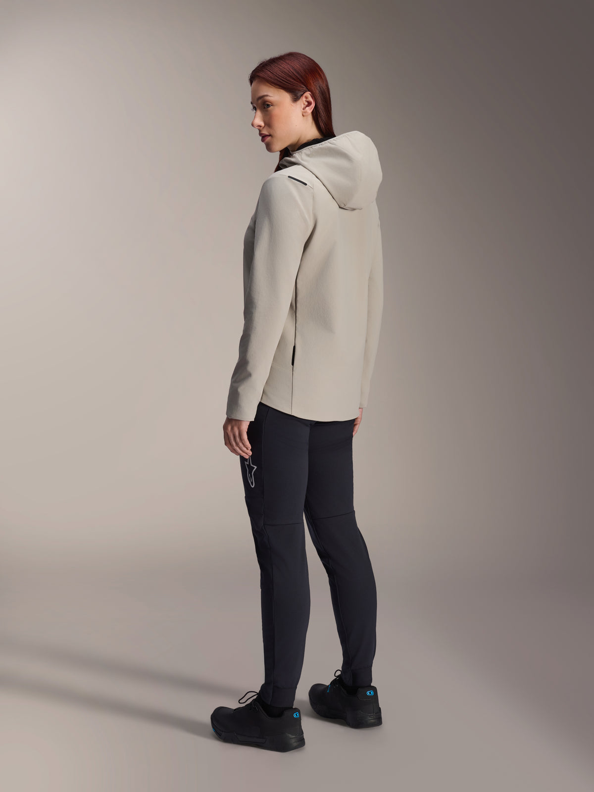 Alpinestars Stella A-Dura Thermal Jacket in Titanium Gray, three-quarter rear view of a woman wearing the insulated hooded cycling jacket with black MTB pants, studio shot showing the regular fit and pocket detail.