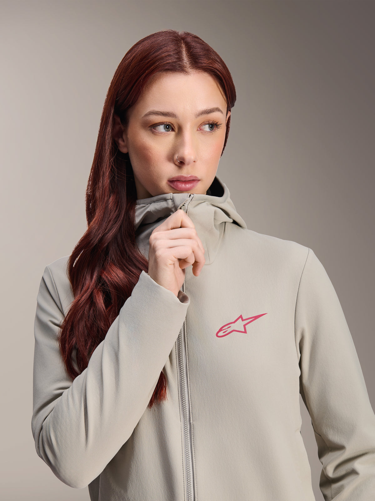 Alpinestars Woman Stella A-Dura Thermal Jacket, Titanium gray with a pink Alpinestars star logo, model with long red hair wearing and zipping up the hooded jacket, lightweight insulated cycling apparel for cold weather.