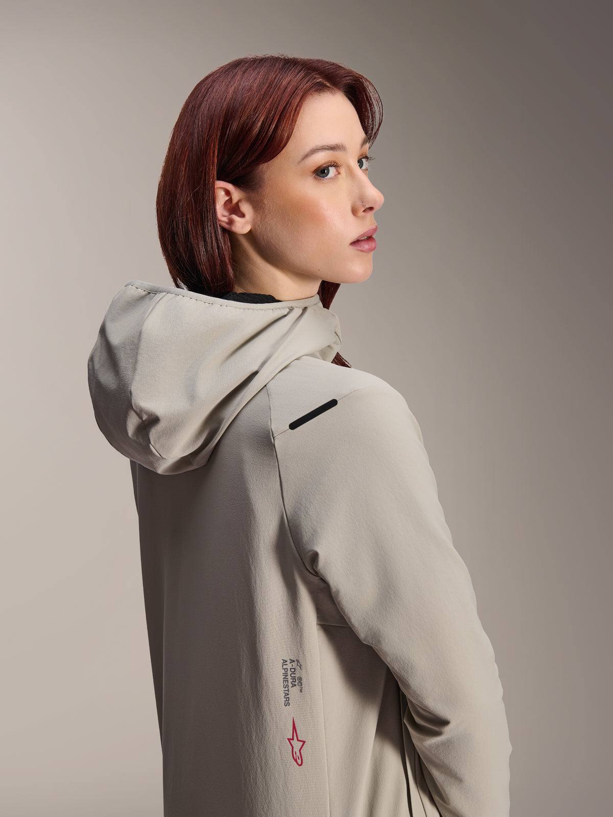 Alpinestars Stella A-Dura Thermal Jacket, Women's MTB Jacket, Titanium Gray, woman looking over her shoulder wearing hooded performance jacket, featuring Alpinestars text and red star logo on the side, designed for cold-weather cycling