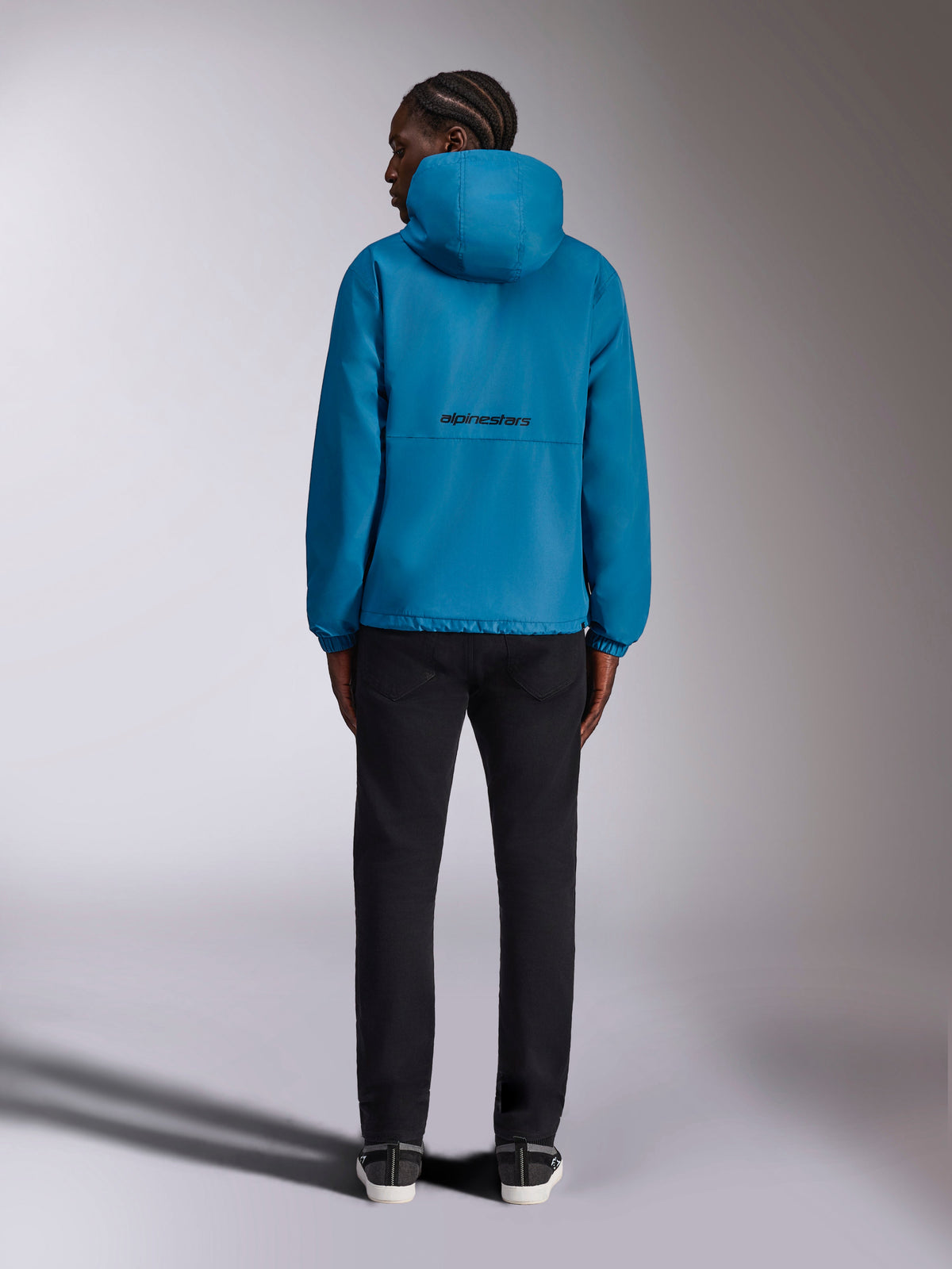 Alpinestars Speeder Chromium Windbreak Jacket, Blue windbreaker, black Alpinestars wordmark logo on the back, hooded design, elastic cuffs, casual sportswear, model seen from the back wearing black pants.