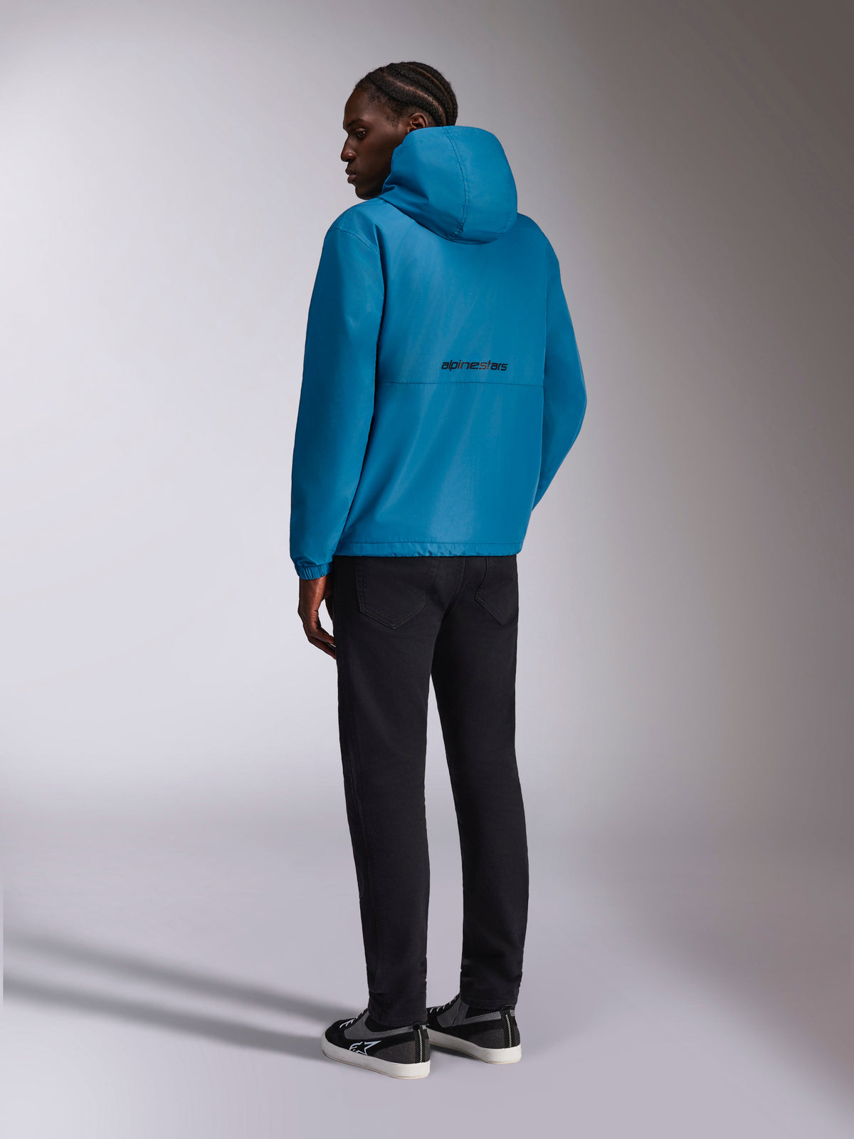 Alpinestars Speeder Chromium Windbreak Jacket, Hooded Windbreaker, Blue, rear view featuring black alpinestars word logo on the back, worn with black pants and black and white sneakers, casual motorsport-inspired streetwear