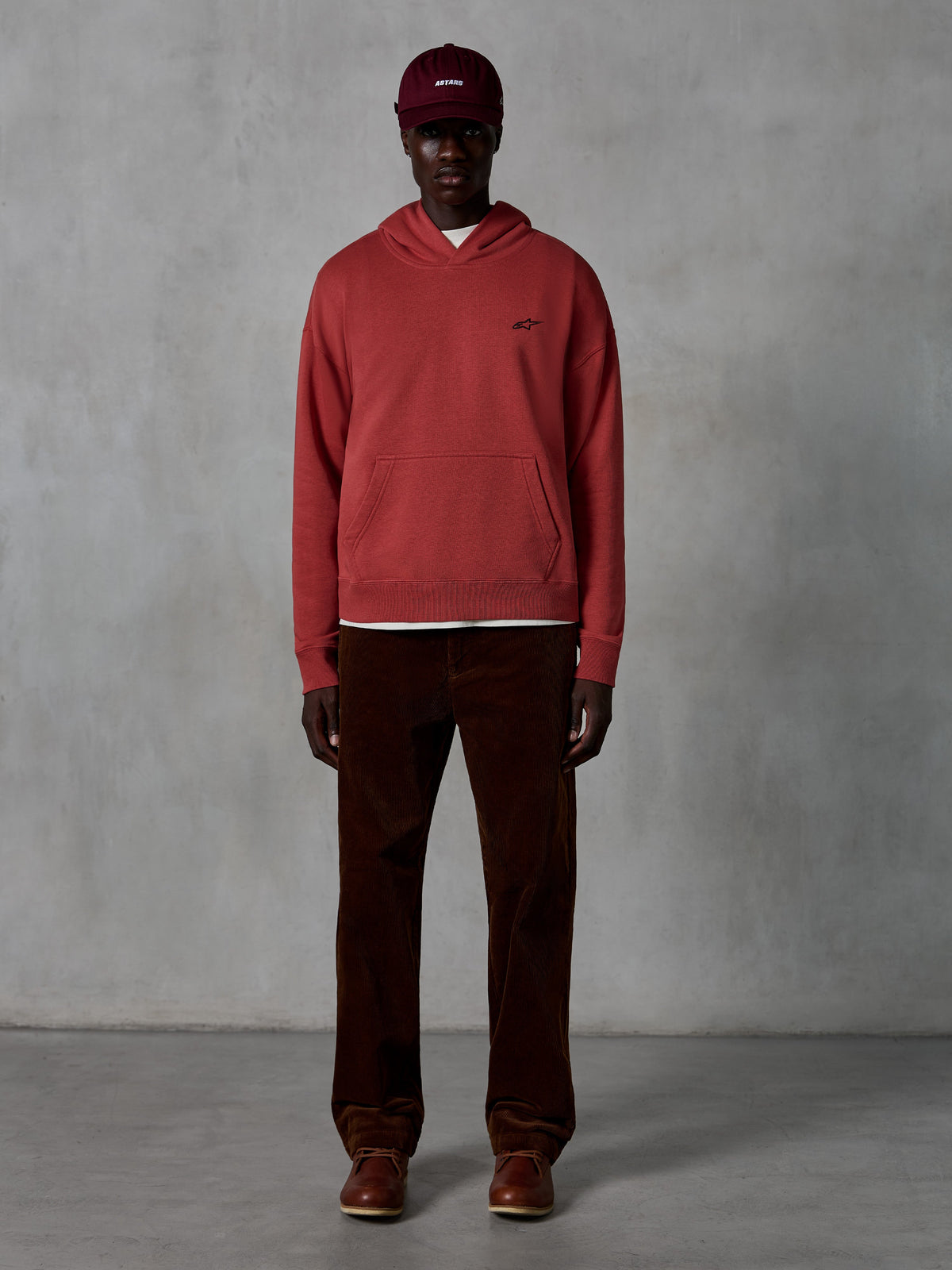 Alpinestars Essentials Ageless Hoodie, casual sportswear hoodie in Brick red with a garment stone wash finish, featuring a small black Alpinestars chest logo and front pouch pocket, worn by a model with a maroon cap and brown corduroy pants for a clean streetwear look.