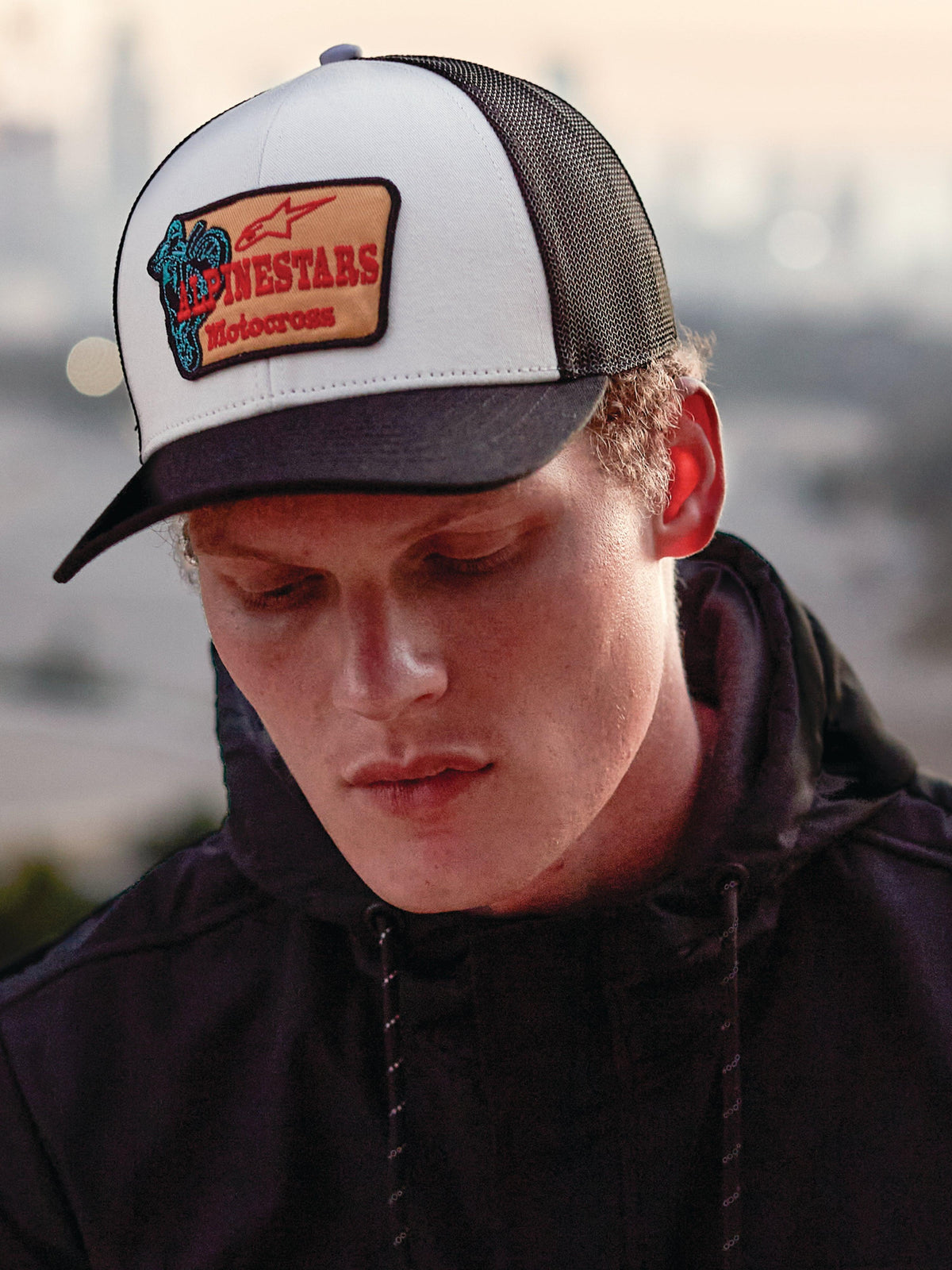 Alpinestars Hardware Trucker Hat in Military White, worn by a man in a black hoodie, featuring a white front panel with a black curved bill and black mesh back, tan embroidered patch with red Alpinestars Motocross branding, casual lifestyle motorsport streetwear