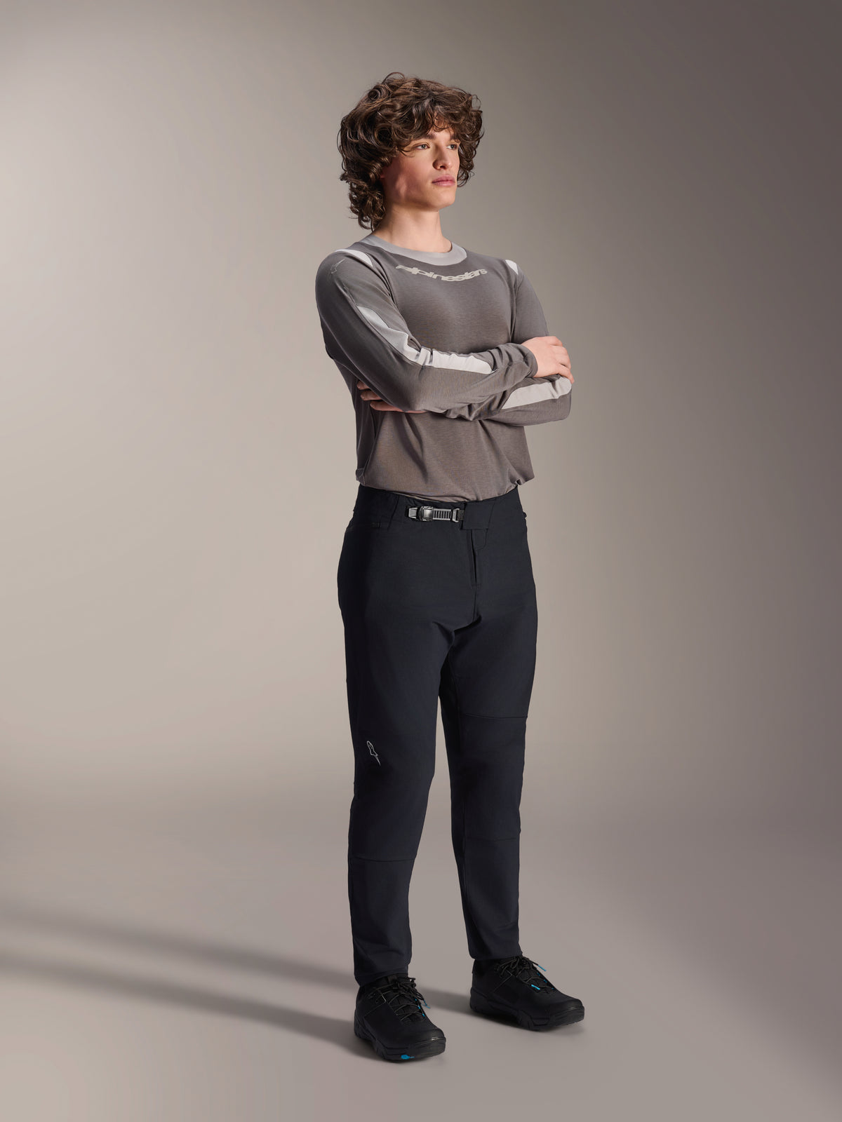 Male model wearing Alpinestars A-Dura Elite Pants, Black MTB Pants, paired with a grey long-sleeve jersey and black cycling shoes, front ratchet closure system, professional mountain bike apparel for trail and enduro riding