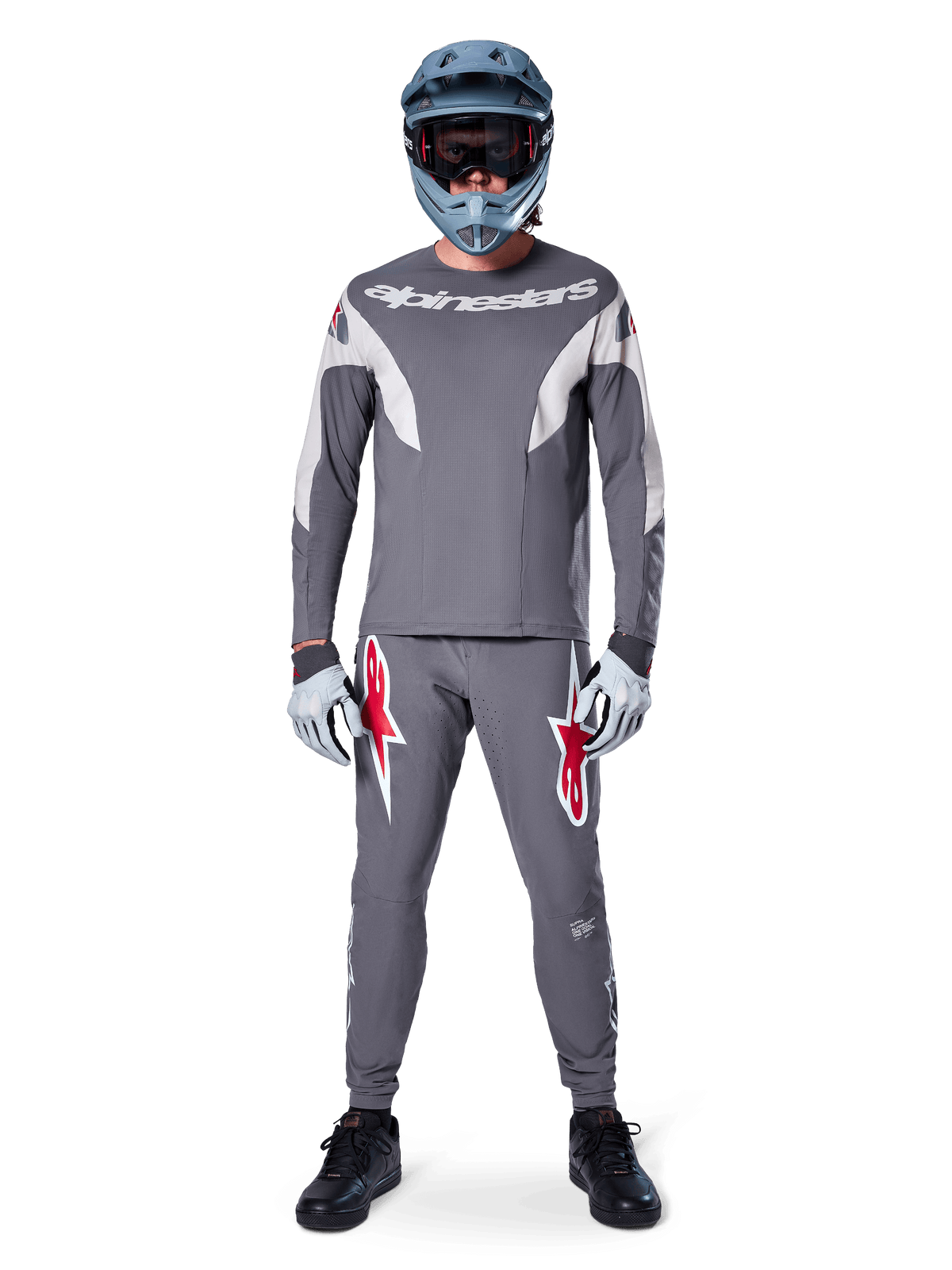 MTB rider wearing Alpinestars A-Supra Race Pants in Dark Gray, featuring bold red logo accents on the thighs, paired with a matching gray jersey, blue full-face helmet, and black shoes for a complete off-road racing kit