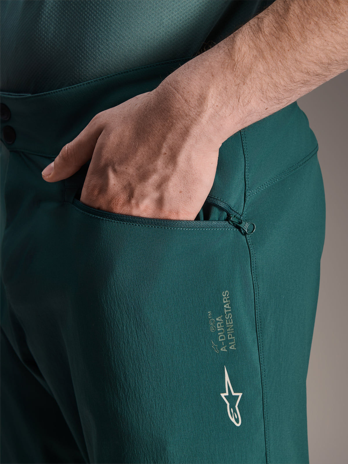 Alpinestars A-Dura Shorts in Dark Pine, close-up view showing a hand in the zippered mesh-lined pocket, featuring white Alpinestars star logo and A-Dura branding, durable textile construction for mountain biking