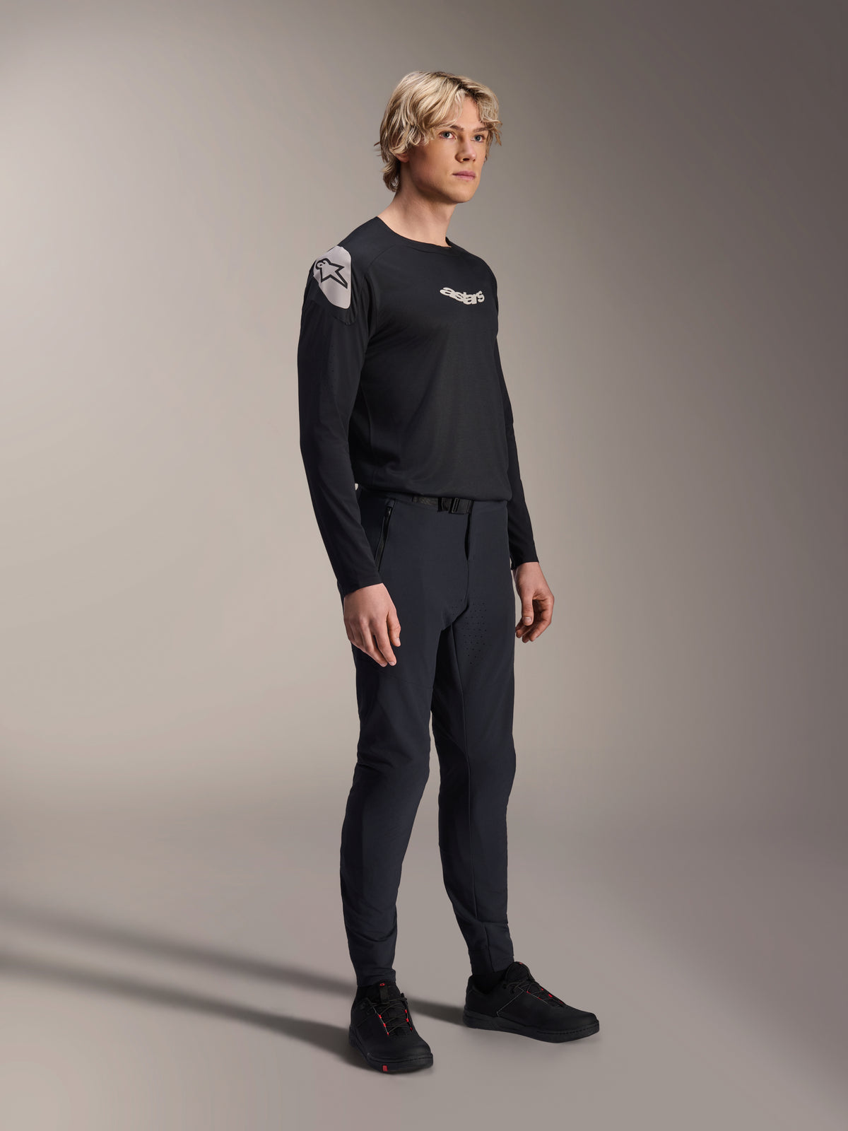 Male model wearing Alpinestars A-Aria Elite Pants in Black Titanium and Aria Polartec Elite LS Jersey, black mountain bike performance gear, featuring laser-perforated ventilation and magnetic buckle closure on the pants, long-sleeve jersey with white logos, complete off-road cycling outfit
