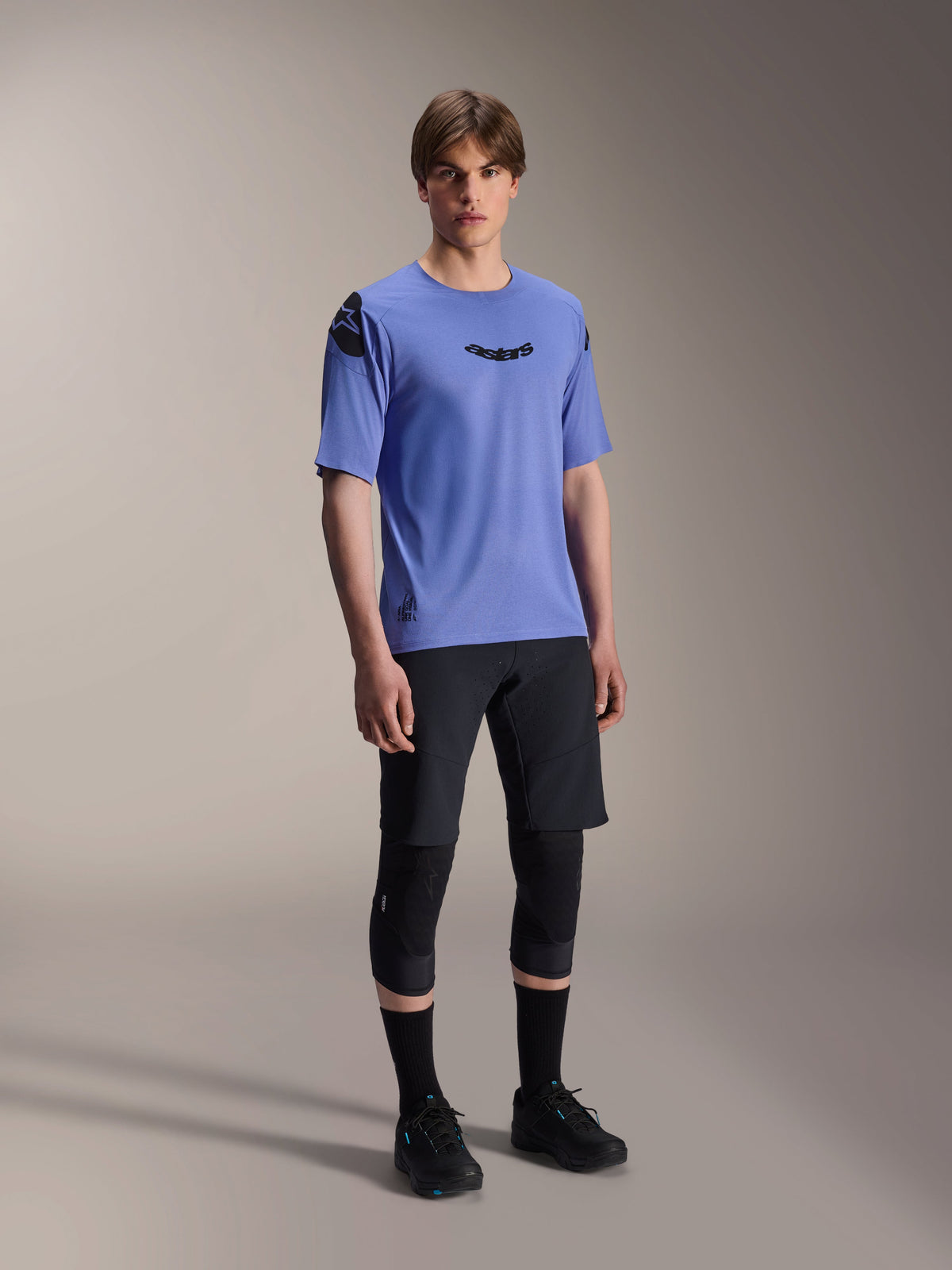Male model wearing Alpinestars A-Aria Polartec Elite Short Sleeve Jersey in Baja Blue, paired with black MTB shorts and knee guards, featuring black Astars logos on the chest and shoulder, premium performance mountain bike gear