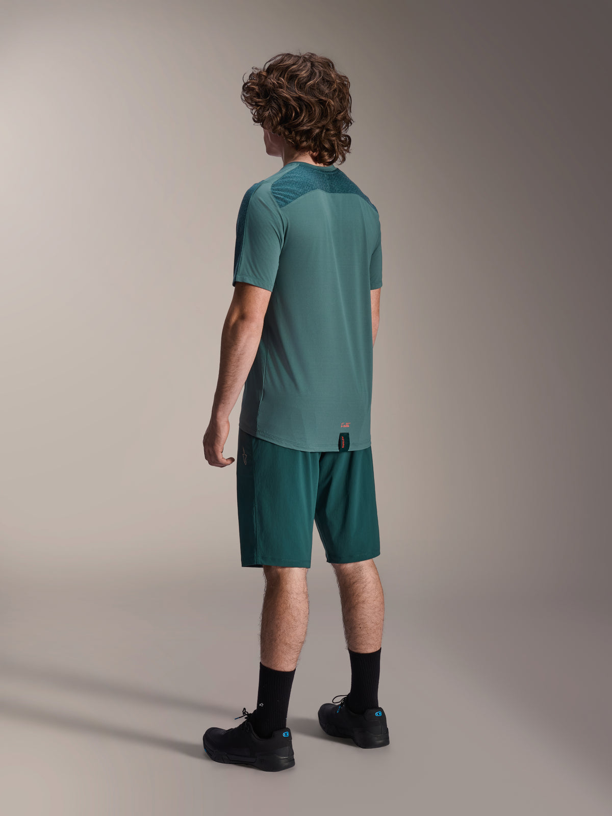 Model wearing Alpinestars A-Dura Shorts in Dark Pine and matching green Lipan short-sleeve jersey, back view, paired with black socks and black mountain bike shoes, professional MTB riding gear.