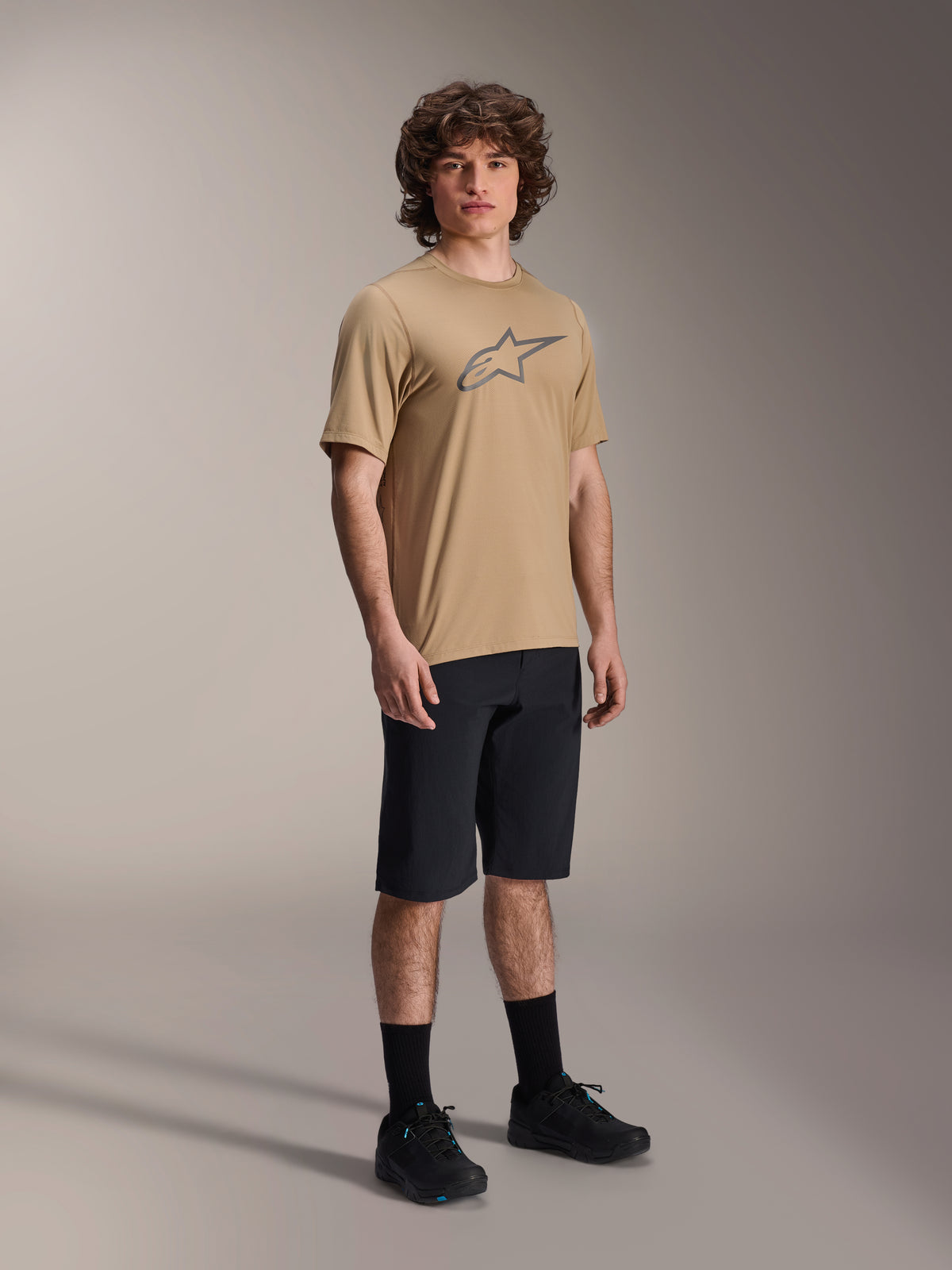 Alpinestars A-Dura Shorts, Black Mountain Bike Shorts, worn by a model with a tan short-sleeve jersey featuring the Alpinestars logo, black socks, and black shoes, durable MTB gear for off-road riding