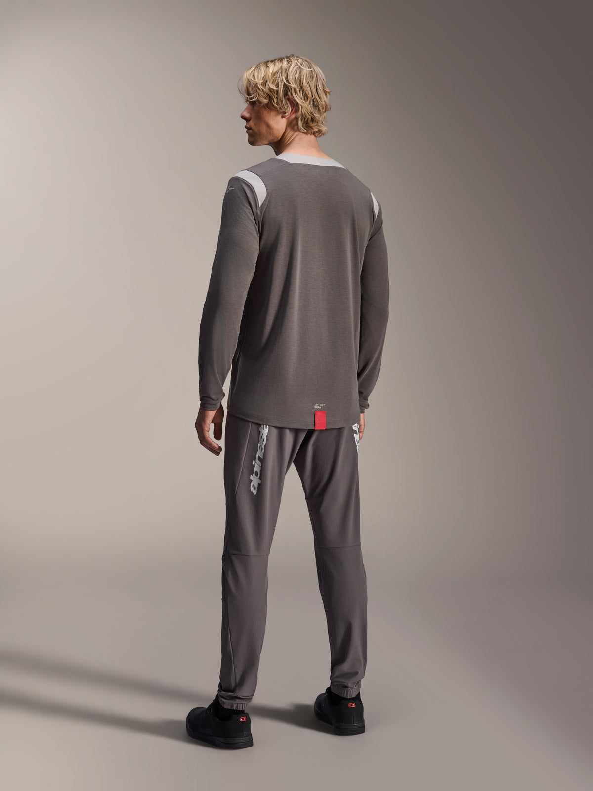 Male model wearing Alpinestars A-Dura Dri Wool Long Sleeve Jersey, Dark Gray with light gray mesh shoulder panels, paired with matching dark gray cycling pants, back view showing the drop tail hem, mountain bike apparel