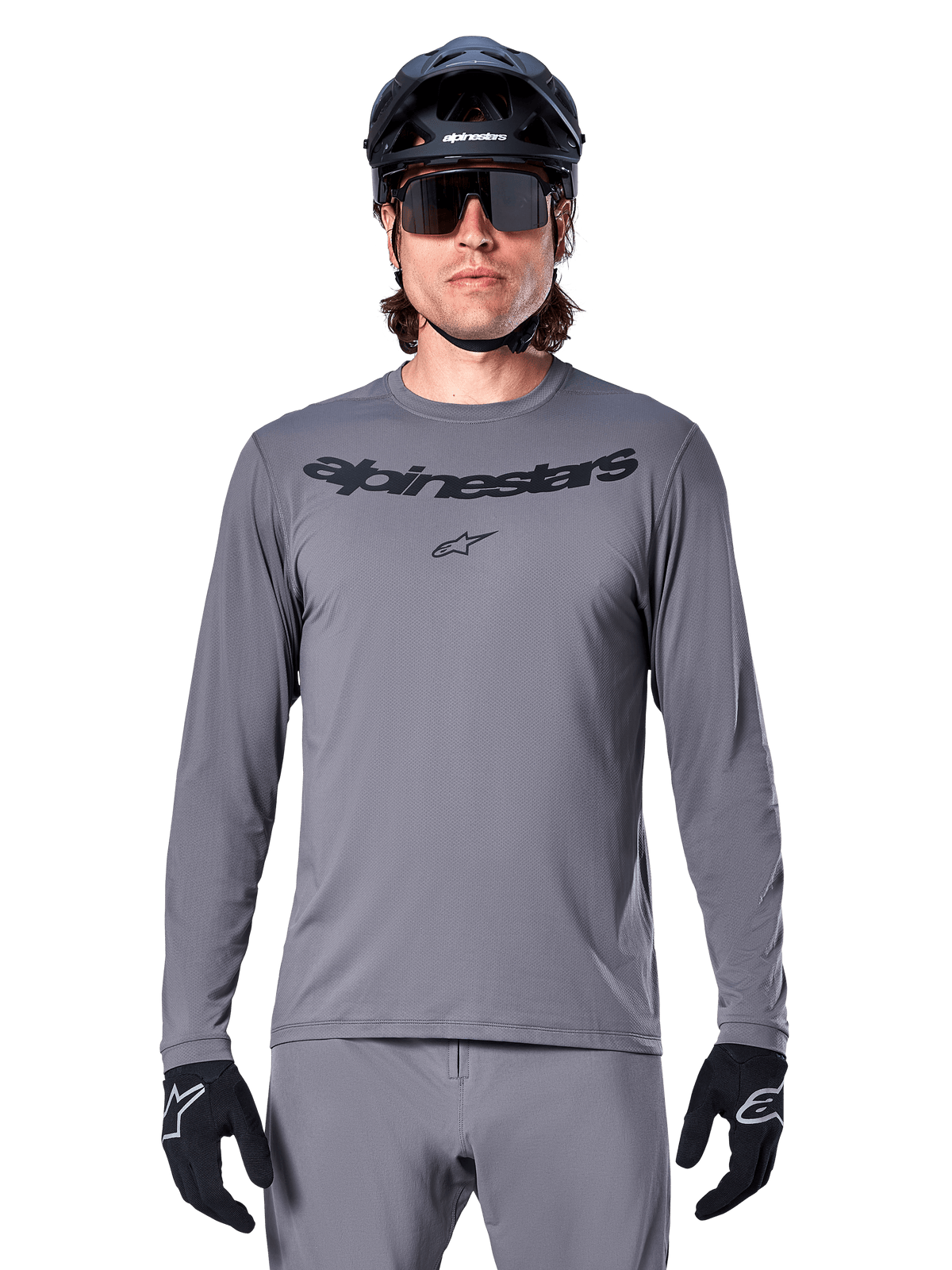 Male rider wearing the Alpinestars A-Dura Rocker Long Sleeve Jersey, Dark Gray MTB jersey with black wordmark on chest, paired with gray pants, black helmet, and gloves, designed for off-road cycling