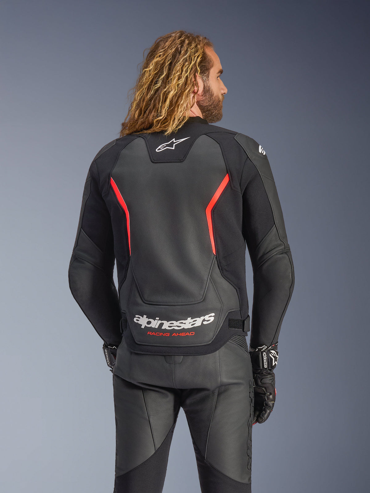 Alpinestars GP Force V2 Leather Jacket, Black and Red Fluo, back view on a model, featuring white Alpinestars logo and 'RACING AHEAD' text, paired with black leather motorcycle pants and gloves.