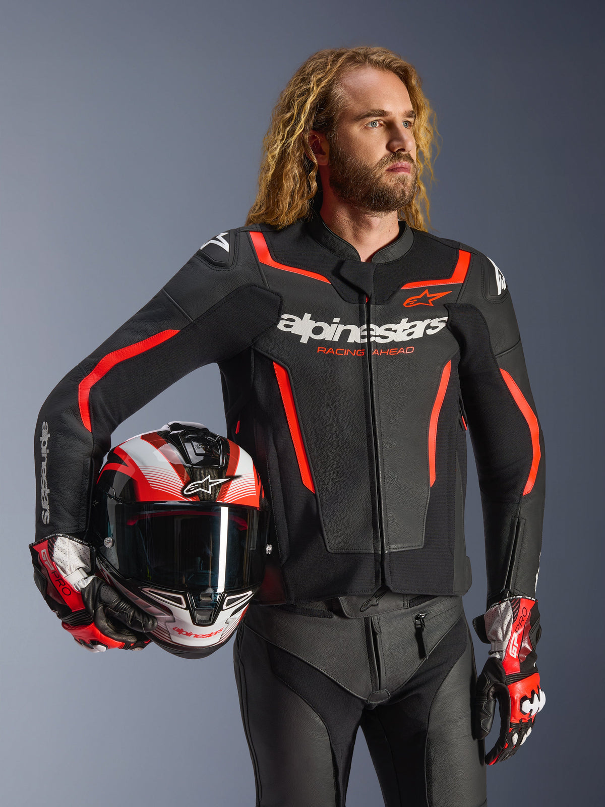 Alpinestars GP Force V2 Leather Jacket, Black and Red Fluo, worn by a male model holding a matching helmet, featuring white Alpinestars logos and red accents, paired with leather pants and racing gloves, sport motorcycle gear for road and track.
