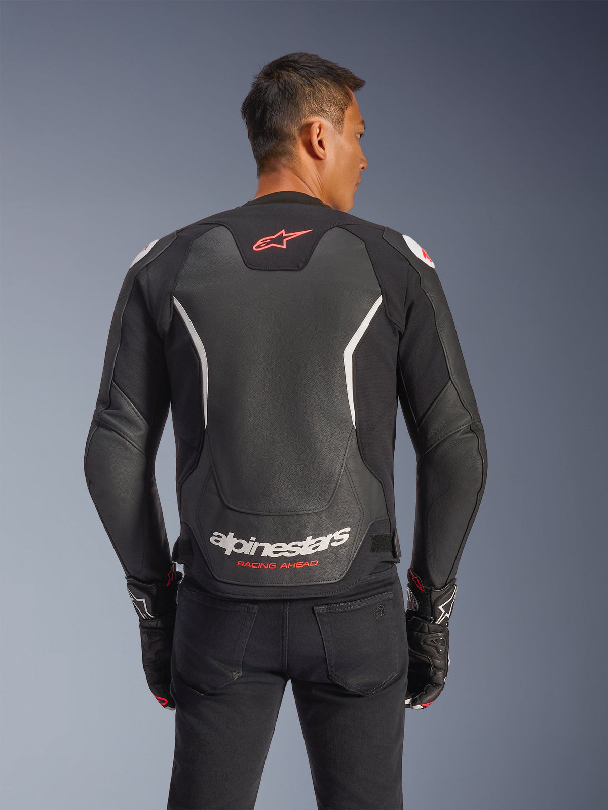 Alpinestars GP Force V2 Leather Jacket, Rear view, Black and White, featuring red star logo on the upper back and white Alpinestars branding with 'Racing Ahead' text, worn with black pants and matching gloves, sport-fit leather motorcycle jacket