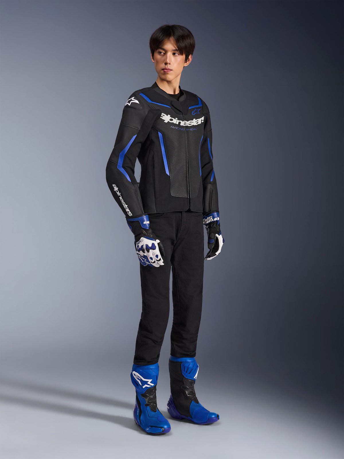Male rider wearing the GP Force V2 Airflow Leather Jacket in Black Metallic Blue, paired with matching racing gloves and blue boots, featuring perforated leather and bold Alpinestars branding, professional motorcycle road gear.