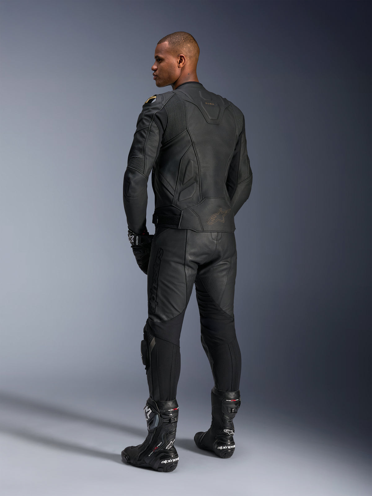 Alpinestars Missile V3 Leather Pants and Dusk Leather Jacket, Black with gold accents, rear view of rider, featuring embossed branding and accordion stretch panels, worn with black Supertech R boots and gloves, professional motorsport apparel.