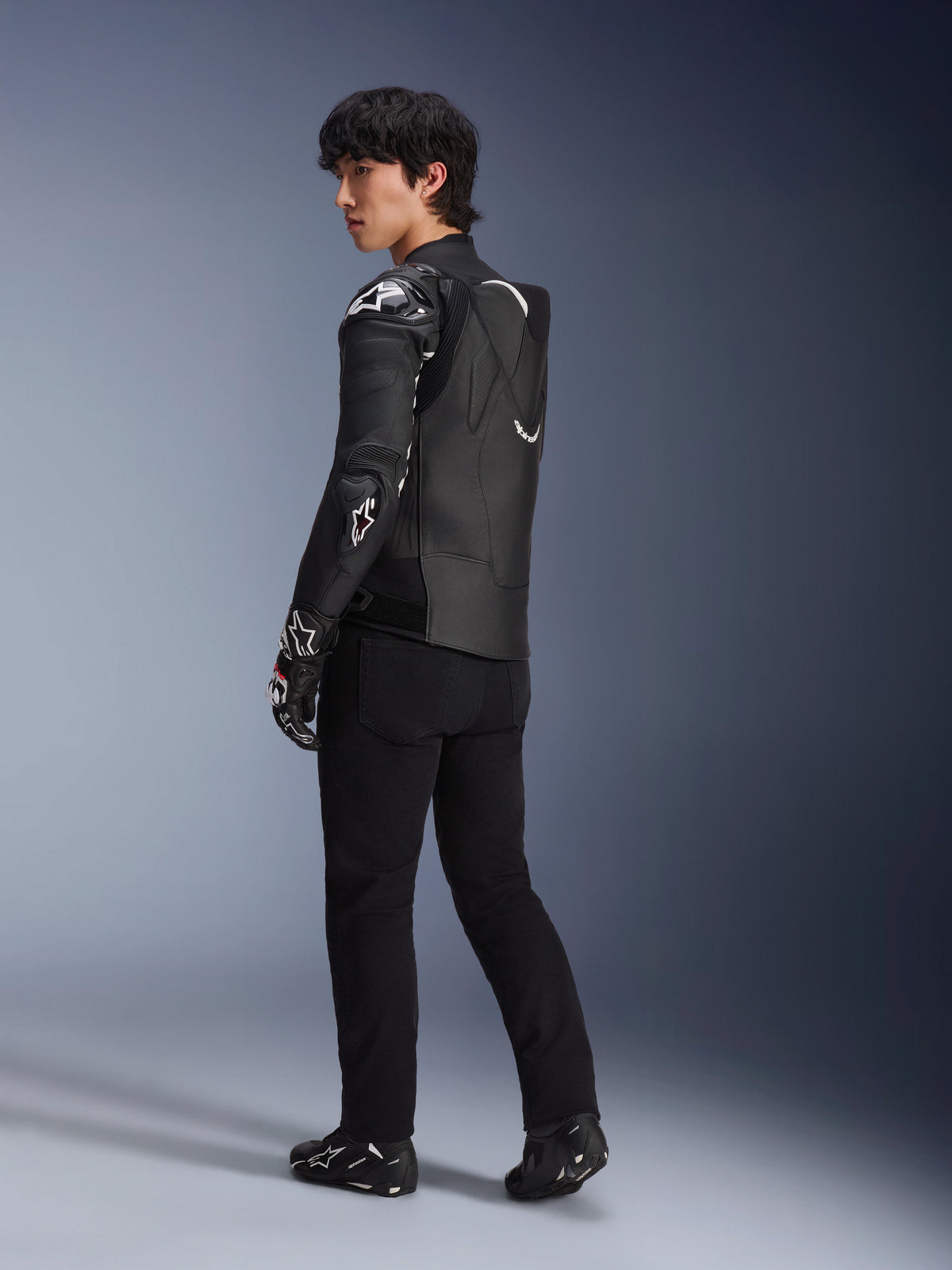 Male model wearing the Alpinestars Atem V5 Leather Jacket in Black and White, three-quarter rear view showing the aerodynamic speed hump and DFS RR protectors, styled with black pants, racing gloves, and boots, professional motorcycle racing apparel