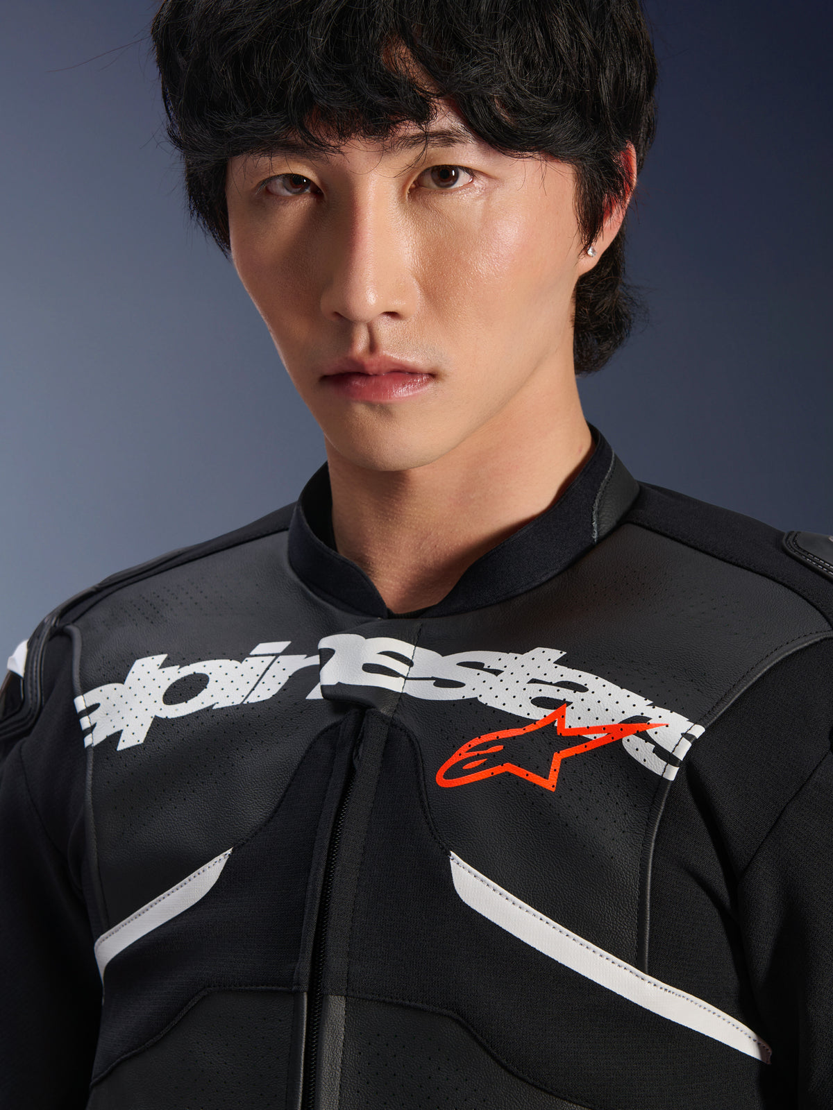 Male model wearing the Alpinestars Atem V5 Leather Jacket in Black and White, close-up shot featuring the large white Alpinestars logo across the chest, red star emblem, perforated leather texture, and technical stretch panels, motorsport lifestyle apparel