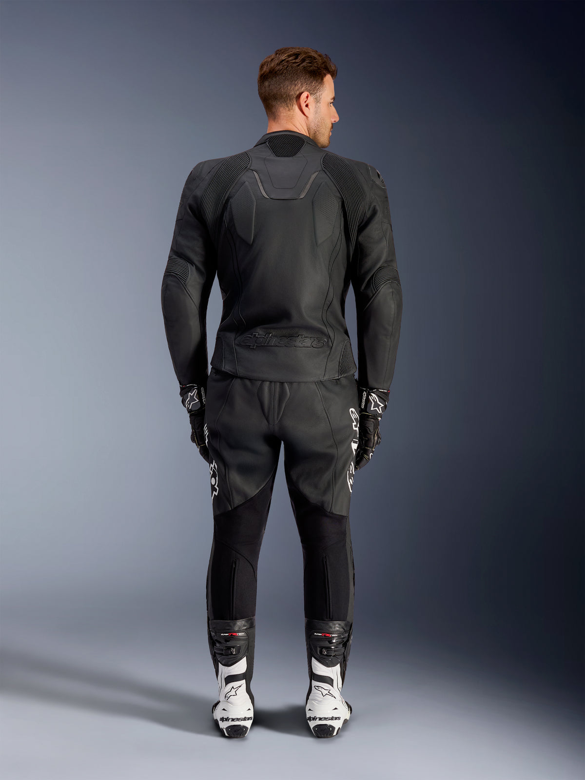 Rear view of a man wearing the Alpinestars Caliber Leather Jacket in Black, featuring an embossed logo and perforated leather panels, paired with black leather motorcycle pants, black gloves, and white racing boots.