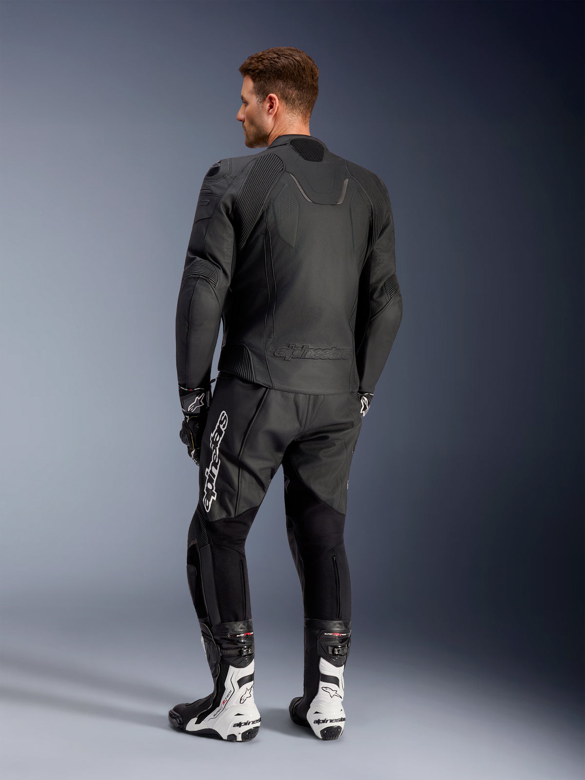 Alpinestars Caliber Leather Jacket, Black motorcycle road jacket, back view, featuring perforated leather and accordion stretch inserts on the waist and shoulders, embossed logo, worn with black leather pants and white racing boots