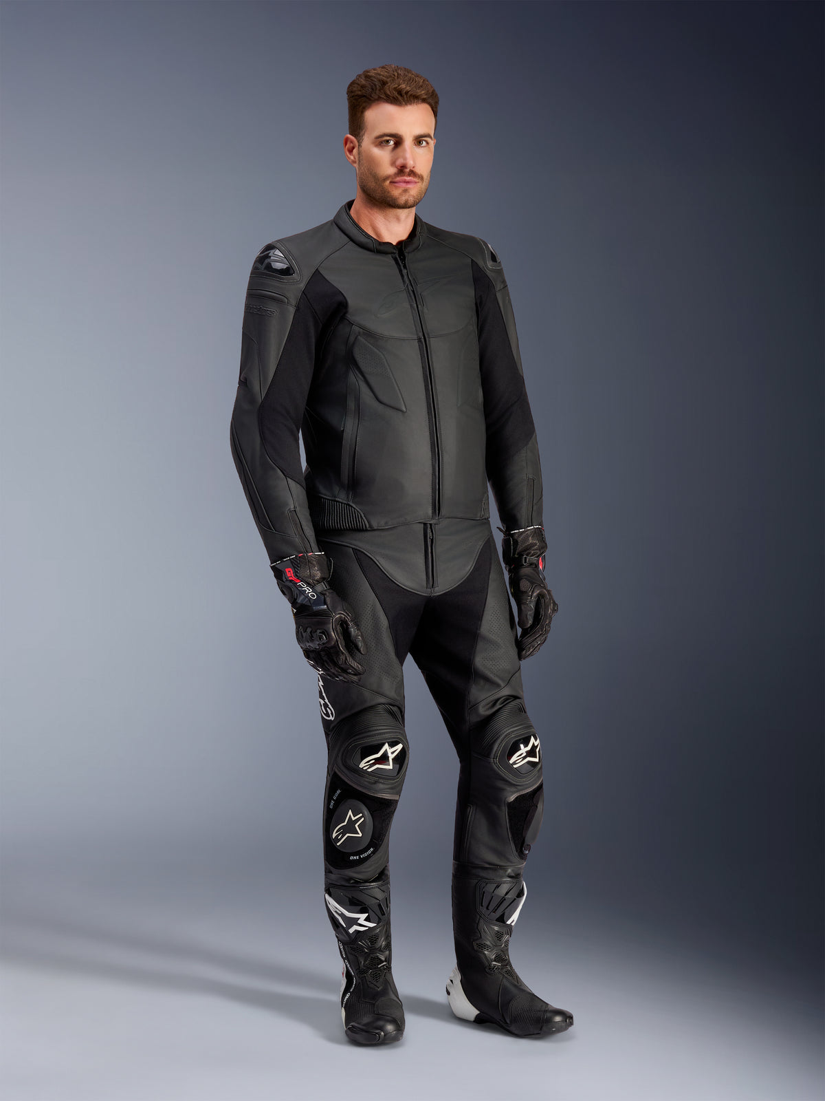 Male model wearing the Alpinestars Caliber Leather Jacket in Black, paired with matching black leather motorcycle pants, racing gloves, and boots, showcasing a complete sport road gear outfit.