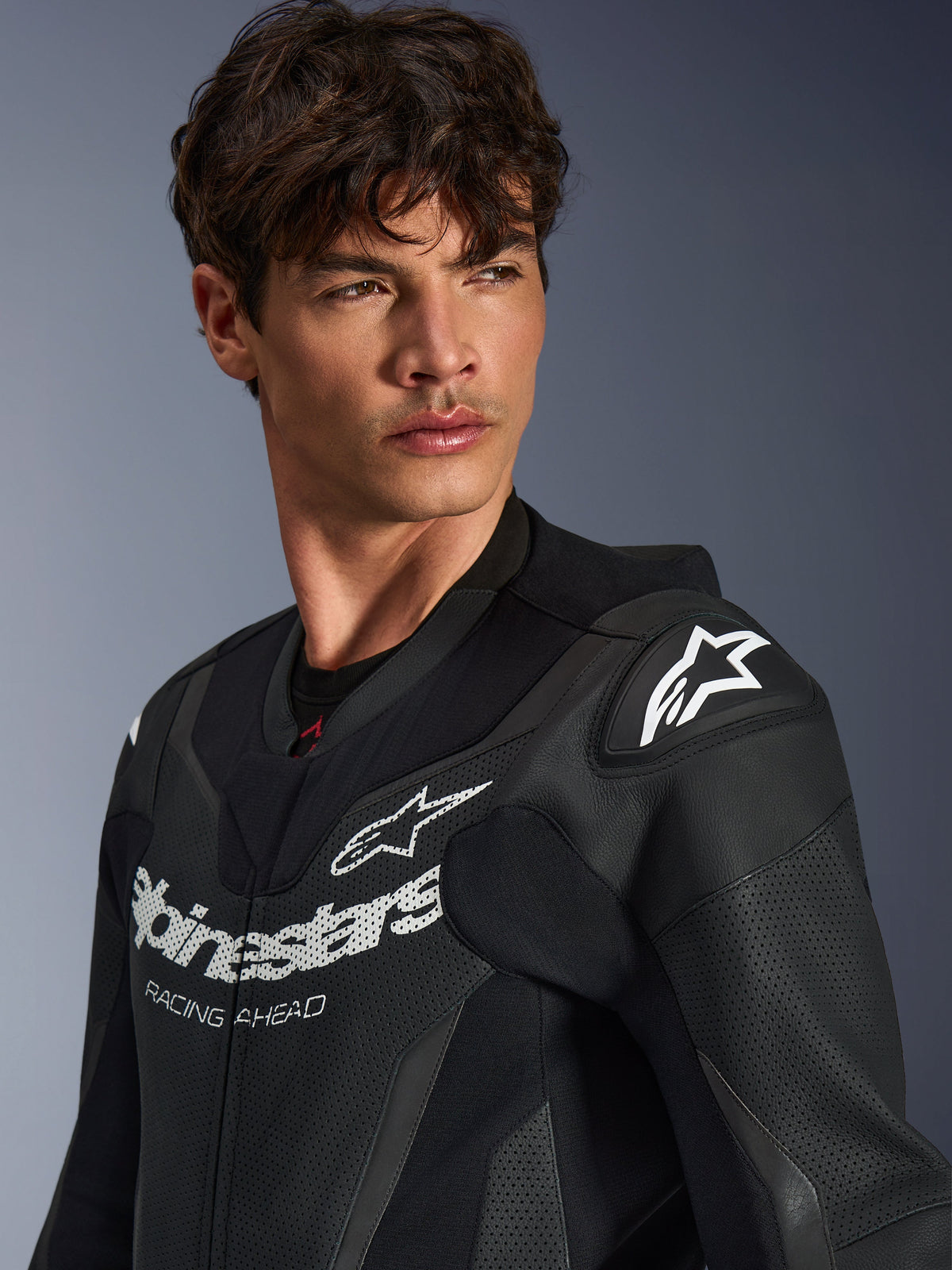 Close-up of a man wearing the Alpinestars GP Force V2 Leather Suit in Black, featuring perforated leather construction, Alpinestars logo with 'RACING AHEAD' branding on the chest, and white star logo on the shoulder sliders.