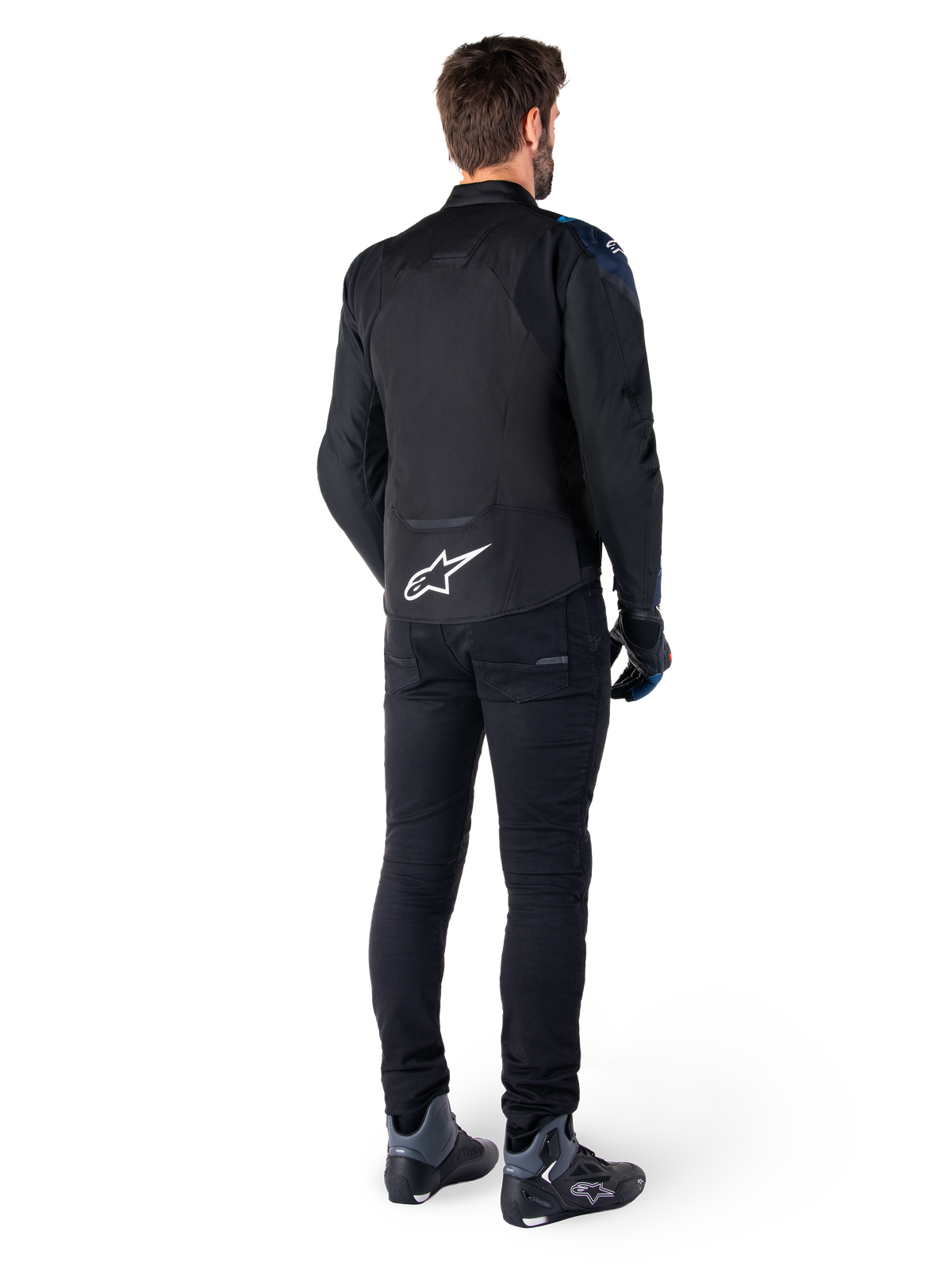 Rear view of a man wearing the Alpinestars T-Jaws V4 Waterproof Jacket in Black, Dark Blue, and Blithe Blue, featuring a prominent white Astar logo on the lower back, paired with black pants and motorcycle riding shoes.