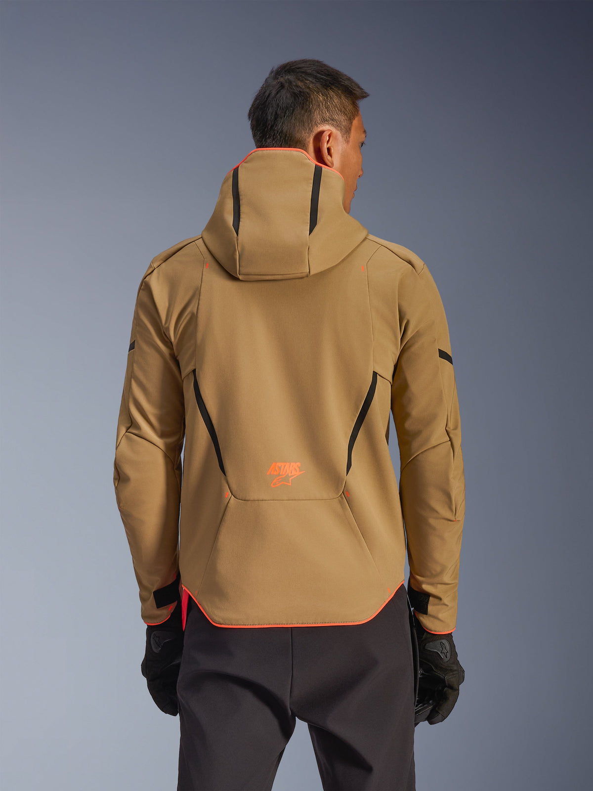 Alpinestars Aeroshell Waterproof Jacket, Dark Khaki and Orange Fluo, rear view showing hoodie design with orange trim on the hood and hem, orange logo on lower back, worn with black gloves, crossover sports softshell motorcycle gear.
