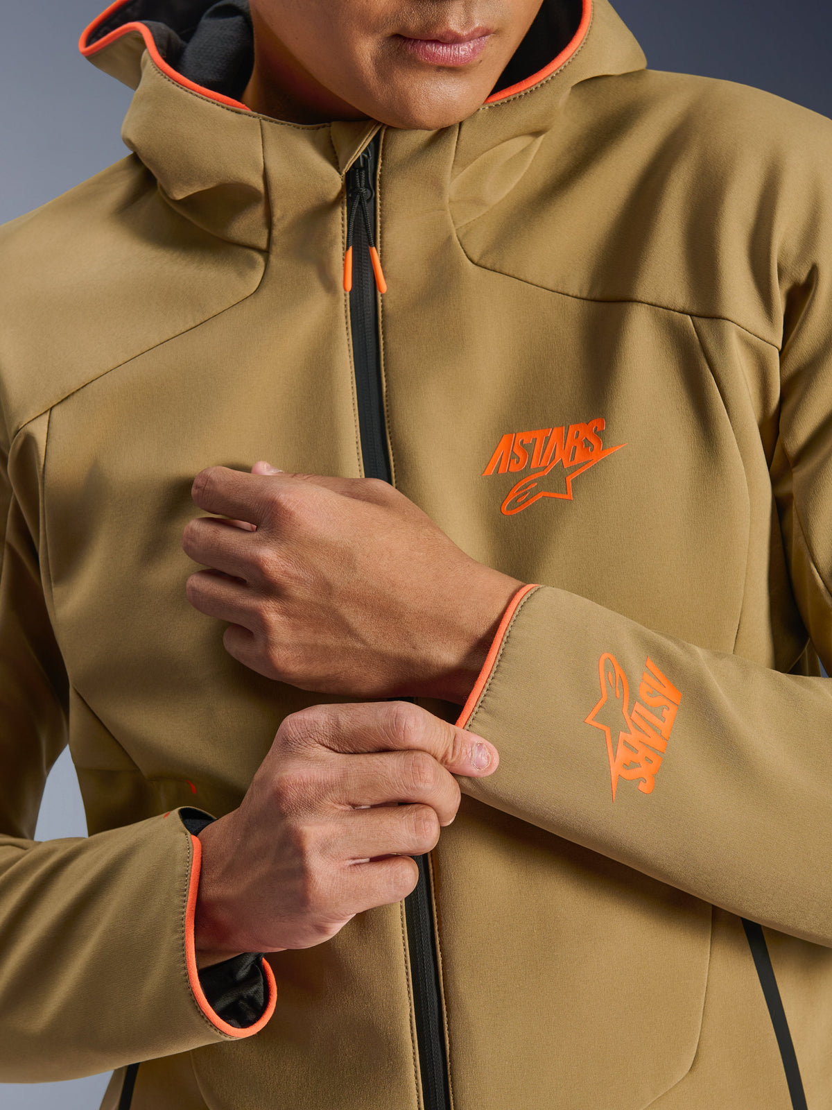 Alpinestars Aeroshell Waterproof Jacket, Dark Khaki and Orange Fluo, close-up of the front and sleeve featuring orange ASTARS logos, orange piping on the hood and cuffs, and a black waterproof zipper.