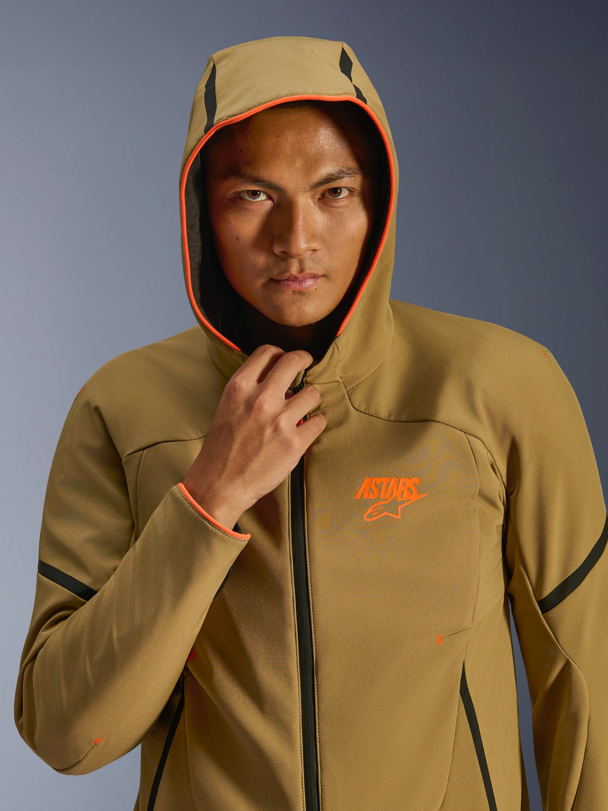Alpinestars Aeroshell Waterproof Jacket, Dark Khaki Black Orange Fluo, model wearing the jacket with the hood up, featuring vibrant orange fluo trim and an orange ASTARS logo, sleek black accents, modern sport-styled waterproof softshell