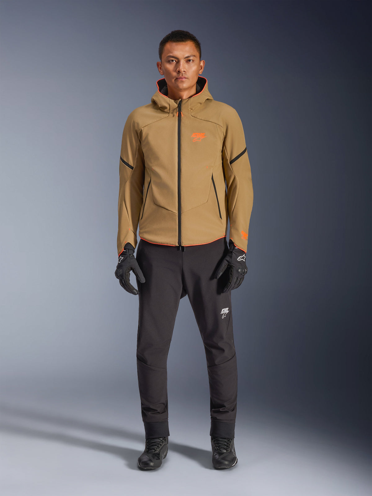 Male model wearing Alpinestars Aeroshell Waterproof Jacket in Dark Khaki with Black and Orange Fluo accents, orange chest logo and hooded design, paired with black pants and black motorcycle gloves, casual sport-touring style