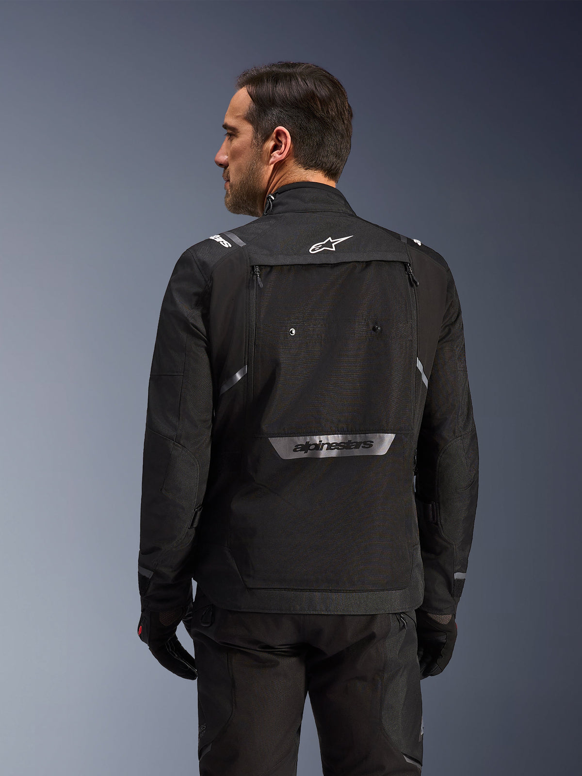 Rider from behind wearing Alpinestars Cusco Drystar® Jacket in Black, textile adventure touring jacket with large rear storage pocket and reflective branding, paired with matching black pants and gloves