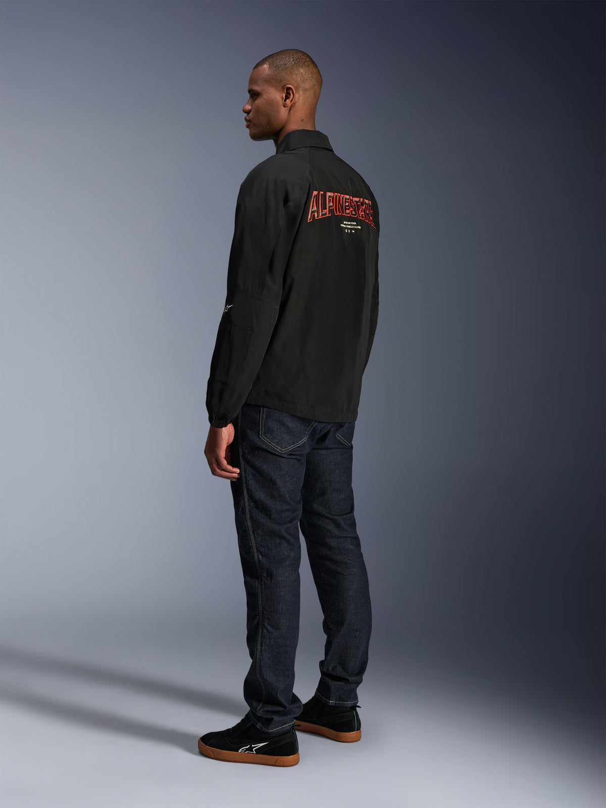 Alpinestars Revved WR Jacket, Black, featuring large orange 3D embroidered Alpinestars branding on the back, rear three-quarter view of a model wearing dark blue jeans and black sneakers, urban contemporary motorcycle jacket with a coaches-jacket silhouette