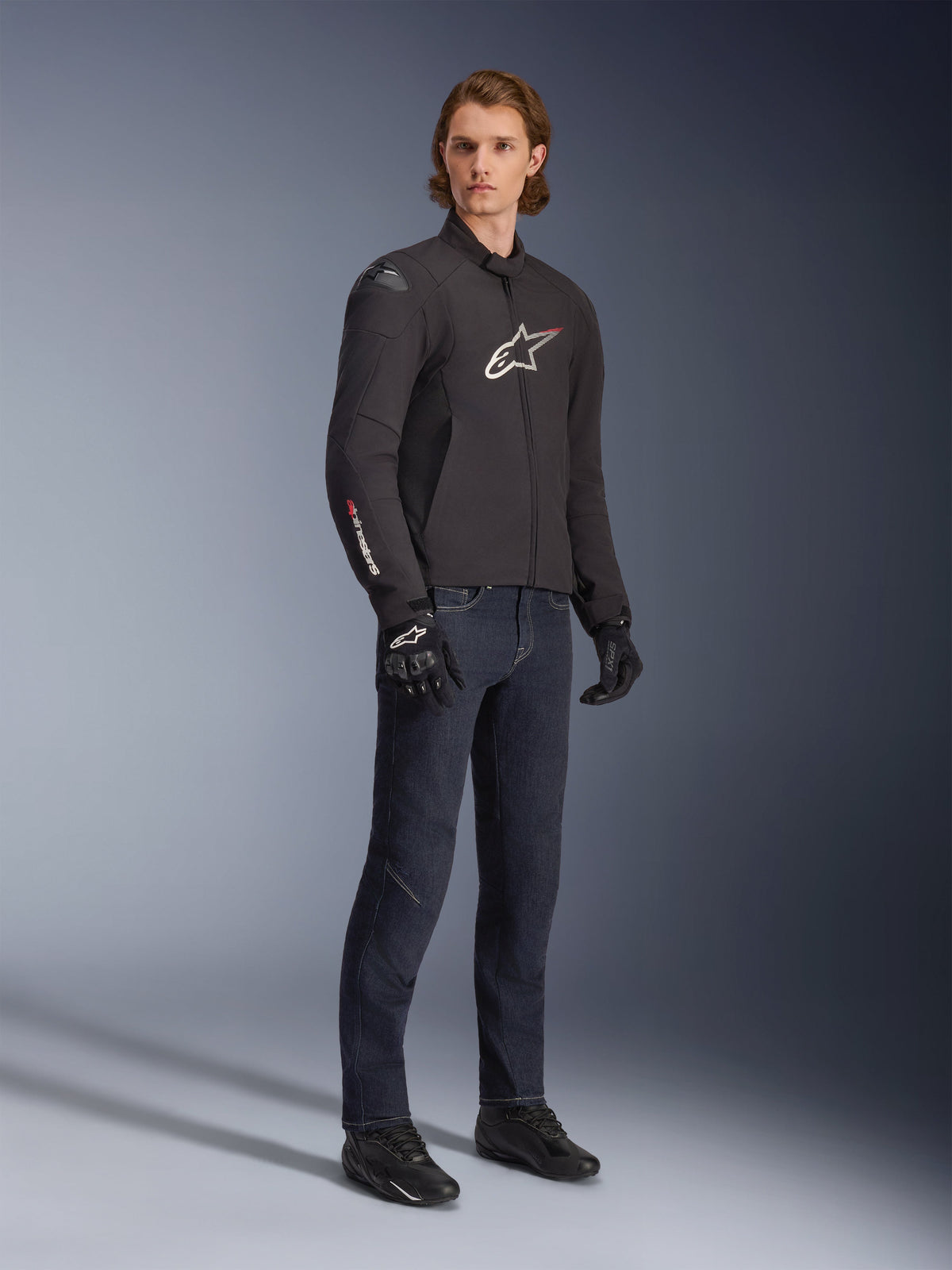 Male model wearing Alpinestars SMX Waterproof Jacket, Black with white and red logo accents, styled with dark blue denim pants and black riding gloves, sleek sport-fit textile motorcycle jacket.