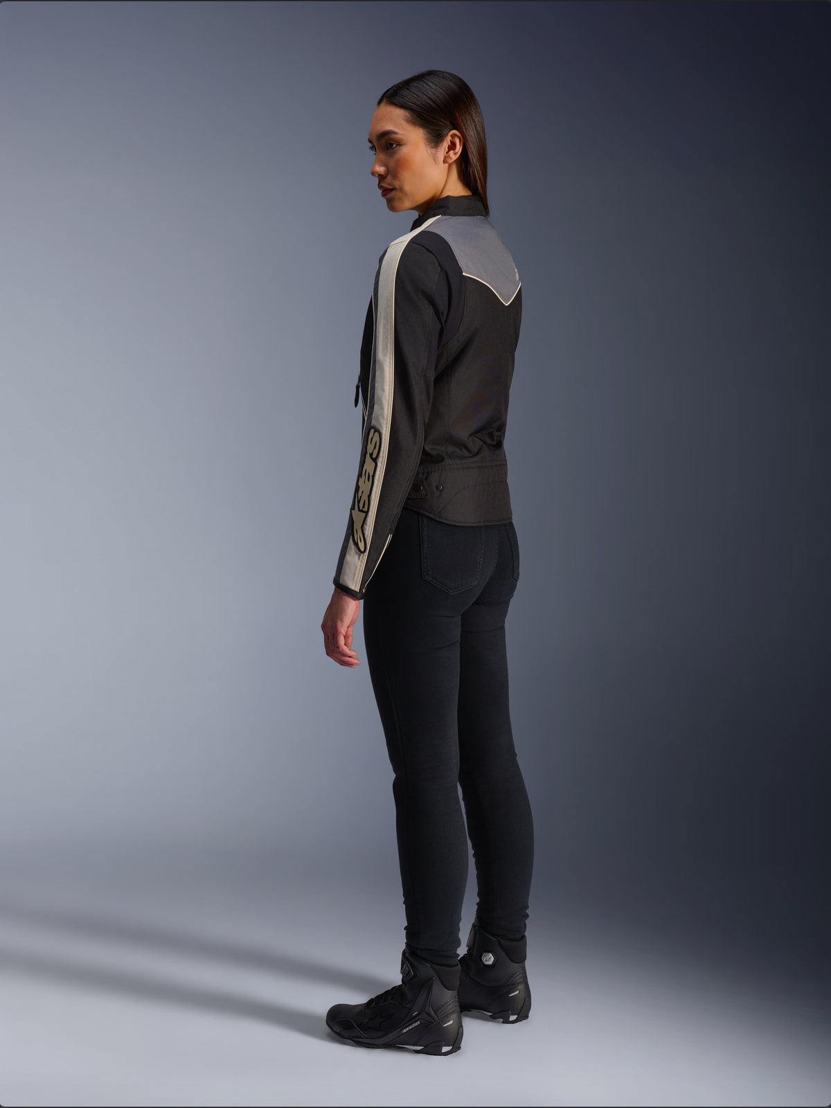 Female model viewed from the side and back wearing the Alpinestars Stella T-Dyno WR Jacket in Black Dark Gray Ecru, featuring vintage-inspired design with ecru sleeve stripes and a gray back panel, worn with black pants and black riding shoes.