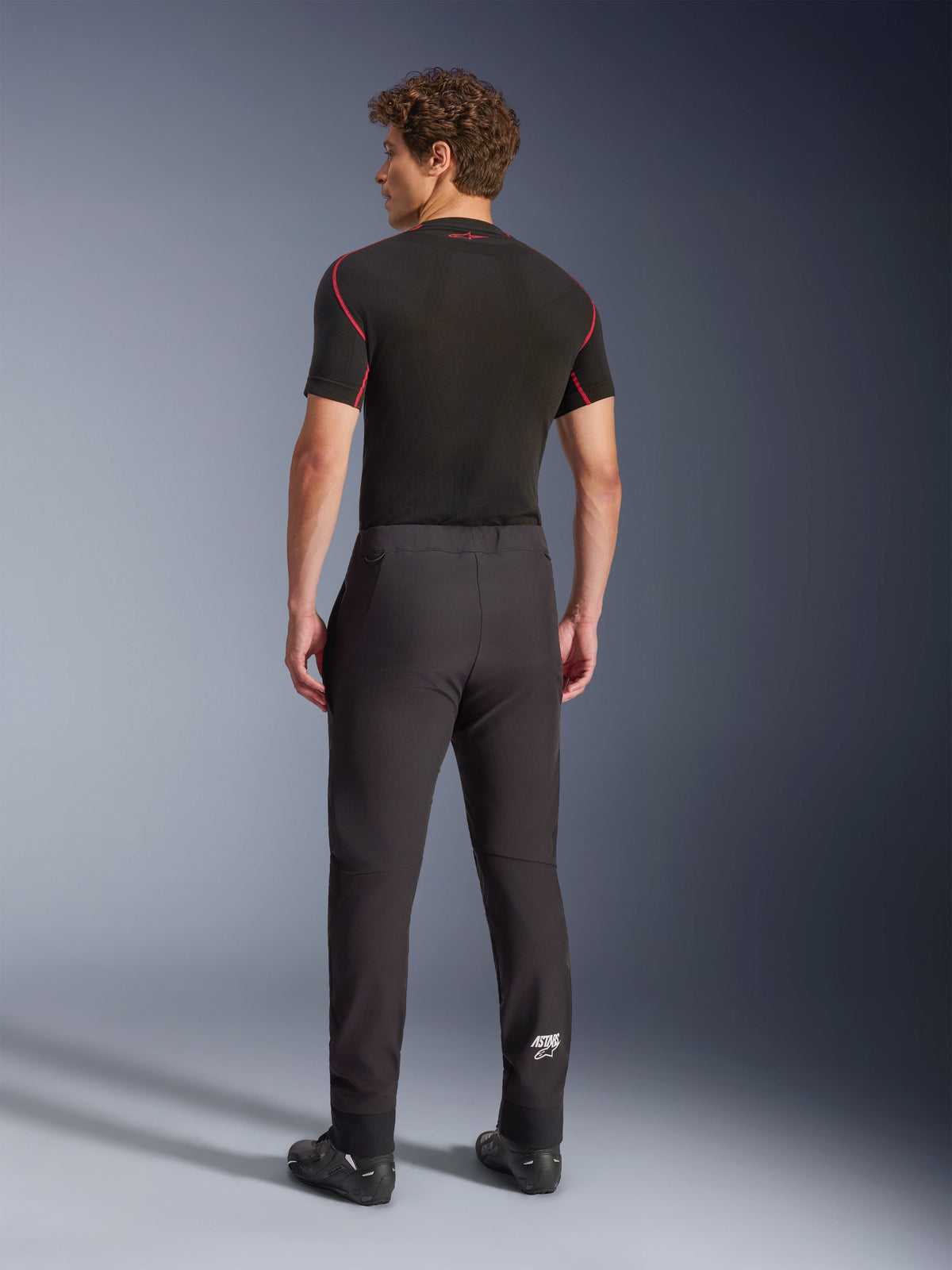 Alpinestars Aeroshell Pants, Black, softshell motorcycle pants, back view on a model, featuring a modern tapered fit, white Astars logo on the lower leg, and elasticated hems, worn with a black short-sleeved base layer and riding shoes.