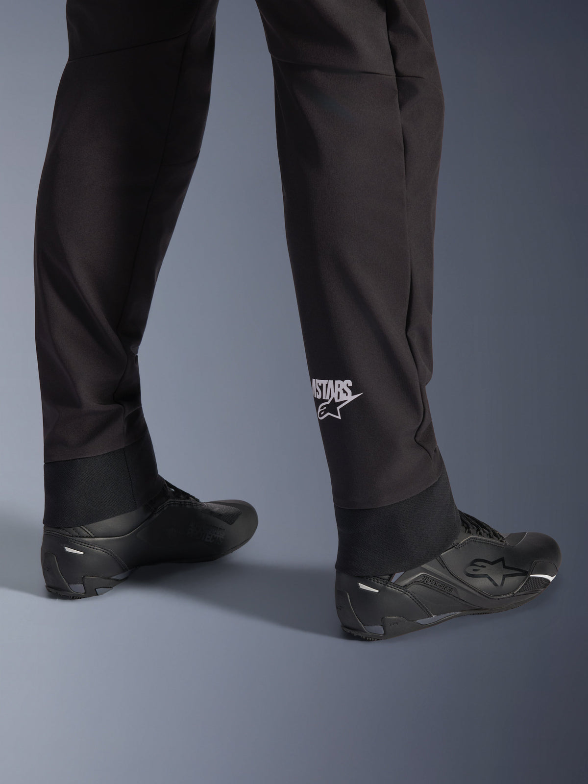 Alpinestars Aeroshell Pants, Black softshell motorcycle pants, close-up of lower leg and elasticated hem, white Alpinestars logo on calf, styled with black riding shoes, modern crossover sport design
