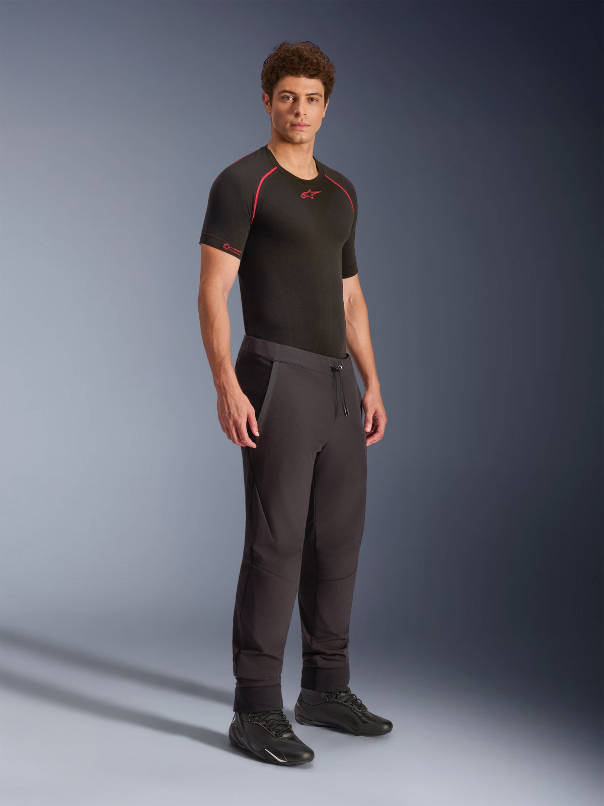 Alpinestars Aeroshell Pants, Black, crossover sports softshell motorcycle pants, worn by a male model with a black performance base layer and black shoes, featuring a drawcord waist and elastic hems.