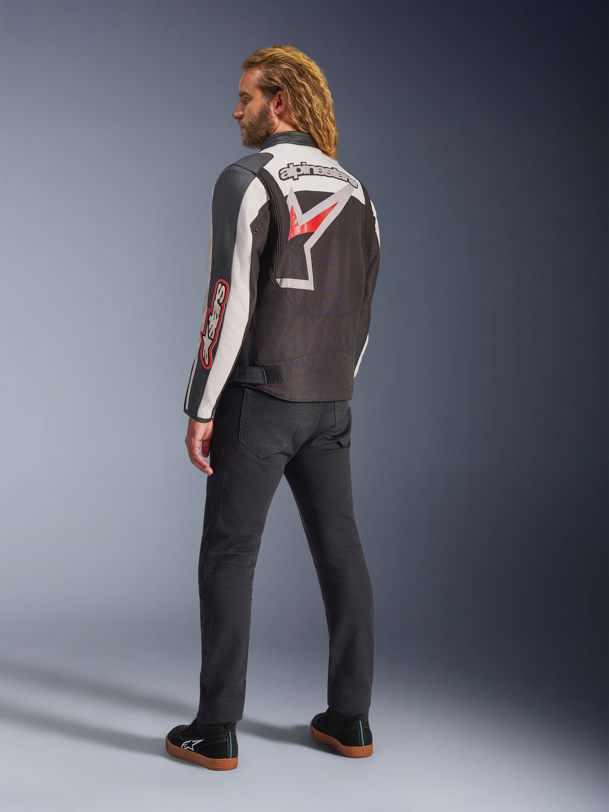 Alpinestars Axton Jacket, Motorcycle Jacket, Black Off White and Bright Red, rear view worn by male model, featuring large Alpinestars star logo and wordmark across the back, vintage racing-inspired design with accordion panels, paired with black pants and sneakers