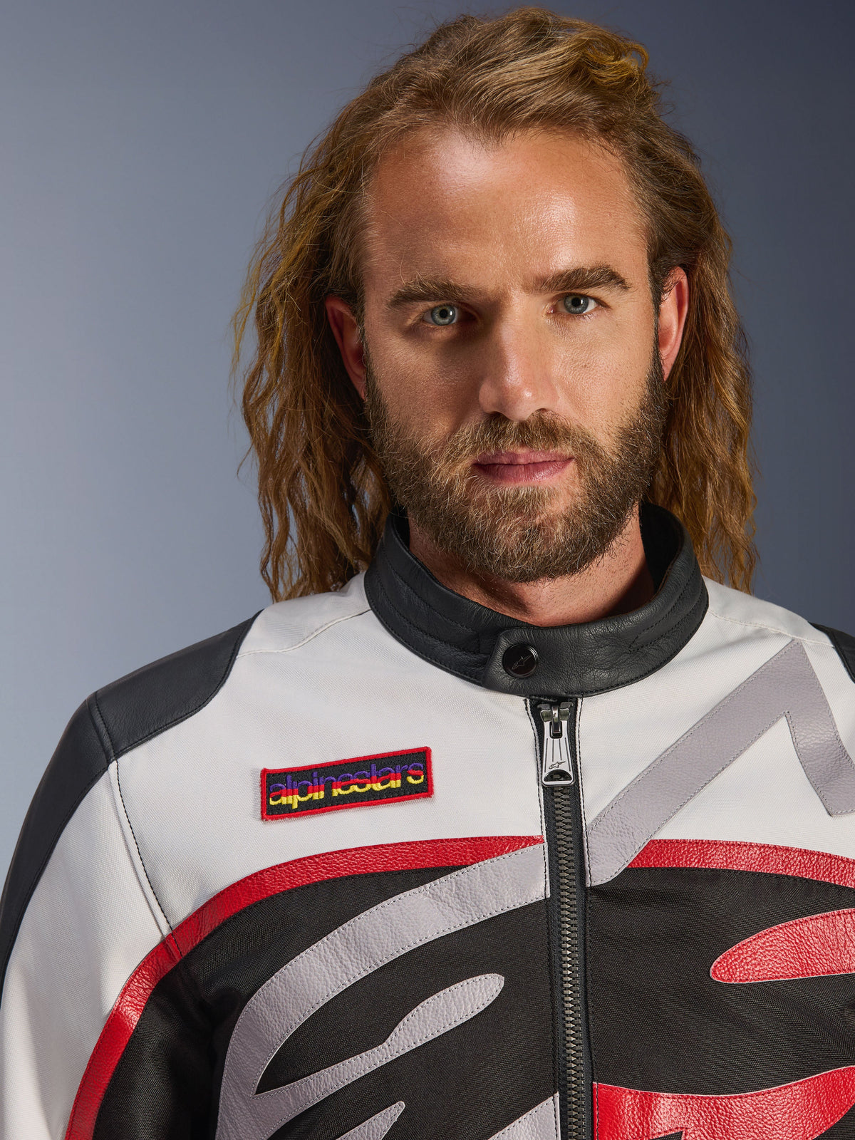 Man wearing the Alpinestars Axton Jacket, vintage-inspired motorcycle jacket in Black Off White and Bright Red, close-up view showing the branded chest patch and minimalist biker collar, contemporary road riding gear