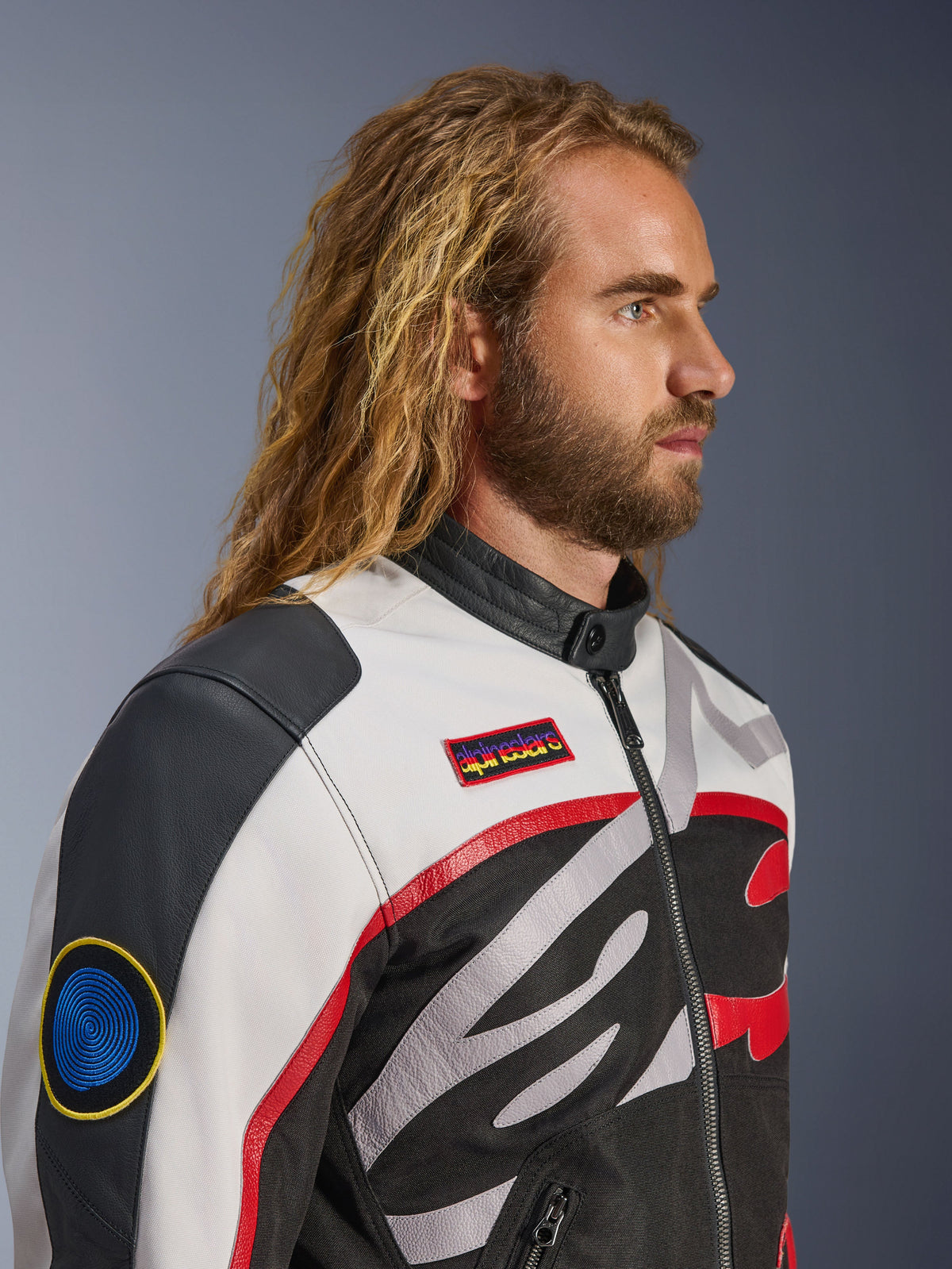 Man with long hair and beard wearing the Alpinestars Axton Jacket, Motorcycle Jacket, Black Off White and Bright Red, vintage racing-inspired design with branded patches and wrap-around logo, close-up profile view, urban contemporary style