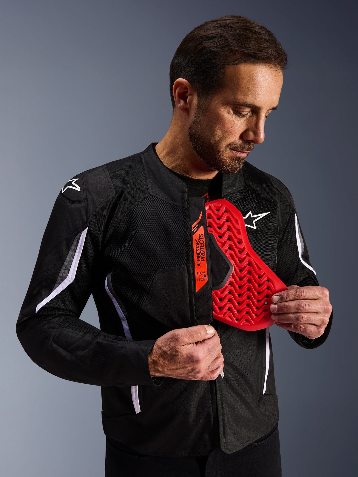 Alpinestars AST V3 Air Jacket in Black, man revealing a bright red honeycomb-patterned chest protector insert, highlighting Tech-Air Ready compatibility and integrated internal armor for motorcycle safety.