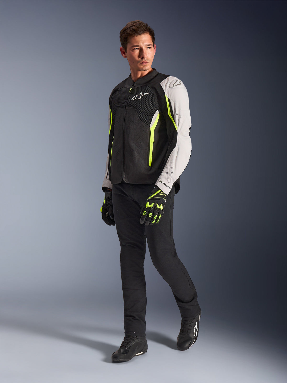 Alpinestars AST V3 Air Jacket, lightweight textile motorcycle jacket in Black, Silver, and Yellow Fluo, worn by a model with black pants and matching gloves, featuring mesh panels and laser perforations for warm-weather sport riding.