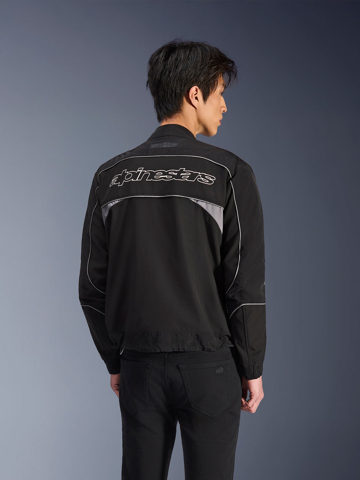 Alpinestars Unite Jacket, Urban Motorcycle Jacket, Black with Dark Gray accents, back view showing white Alpinestars wordmark across the shoulders, reflective piping and panels, minimalist softshell streetwear design